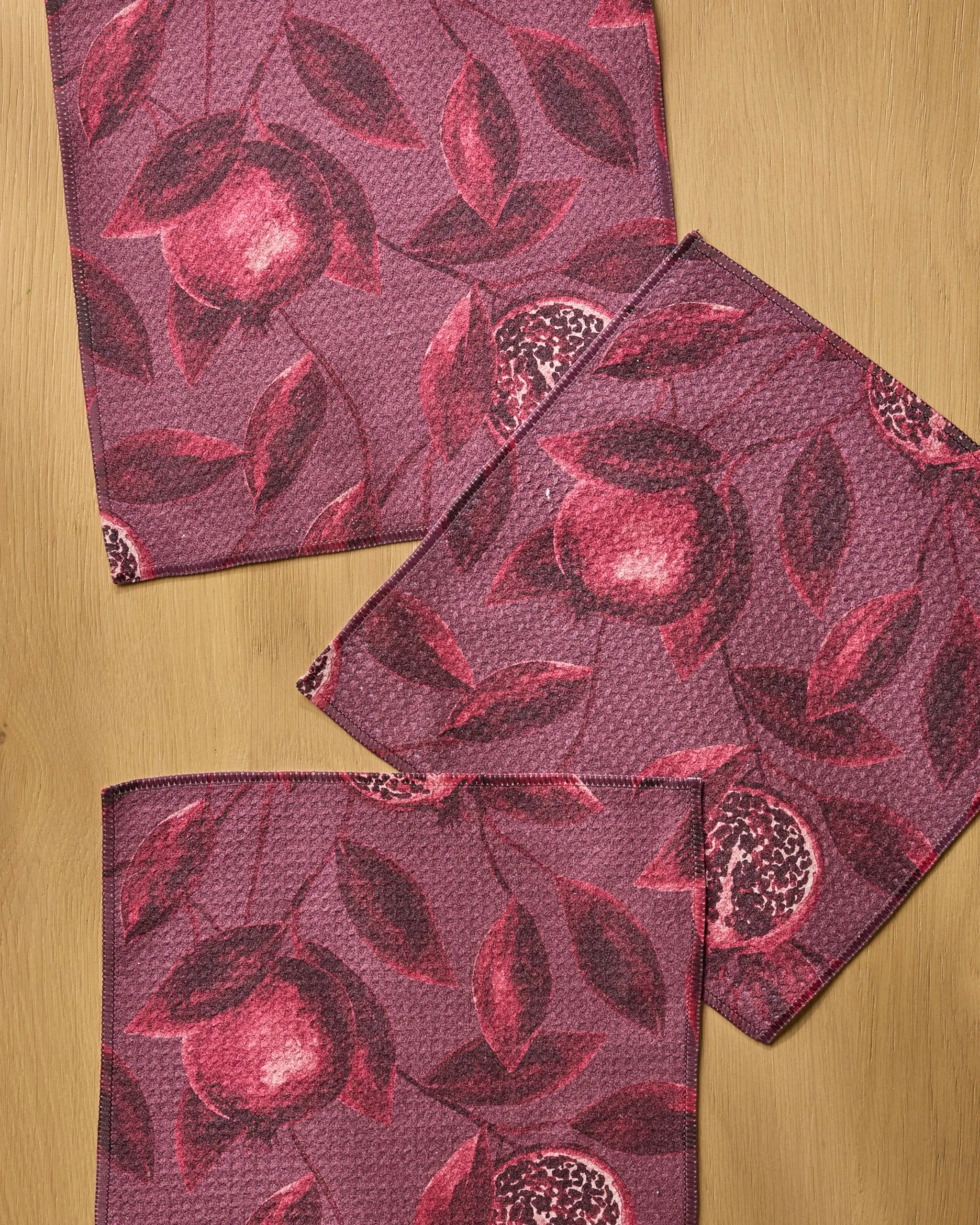 Three "Rich Garnet" napkins by Geometry, crafted from recycled materials and featuring a fruit and leaf pattern in pink and burgundy, are arranged with slight overlap on a light wooden surface.