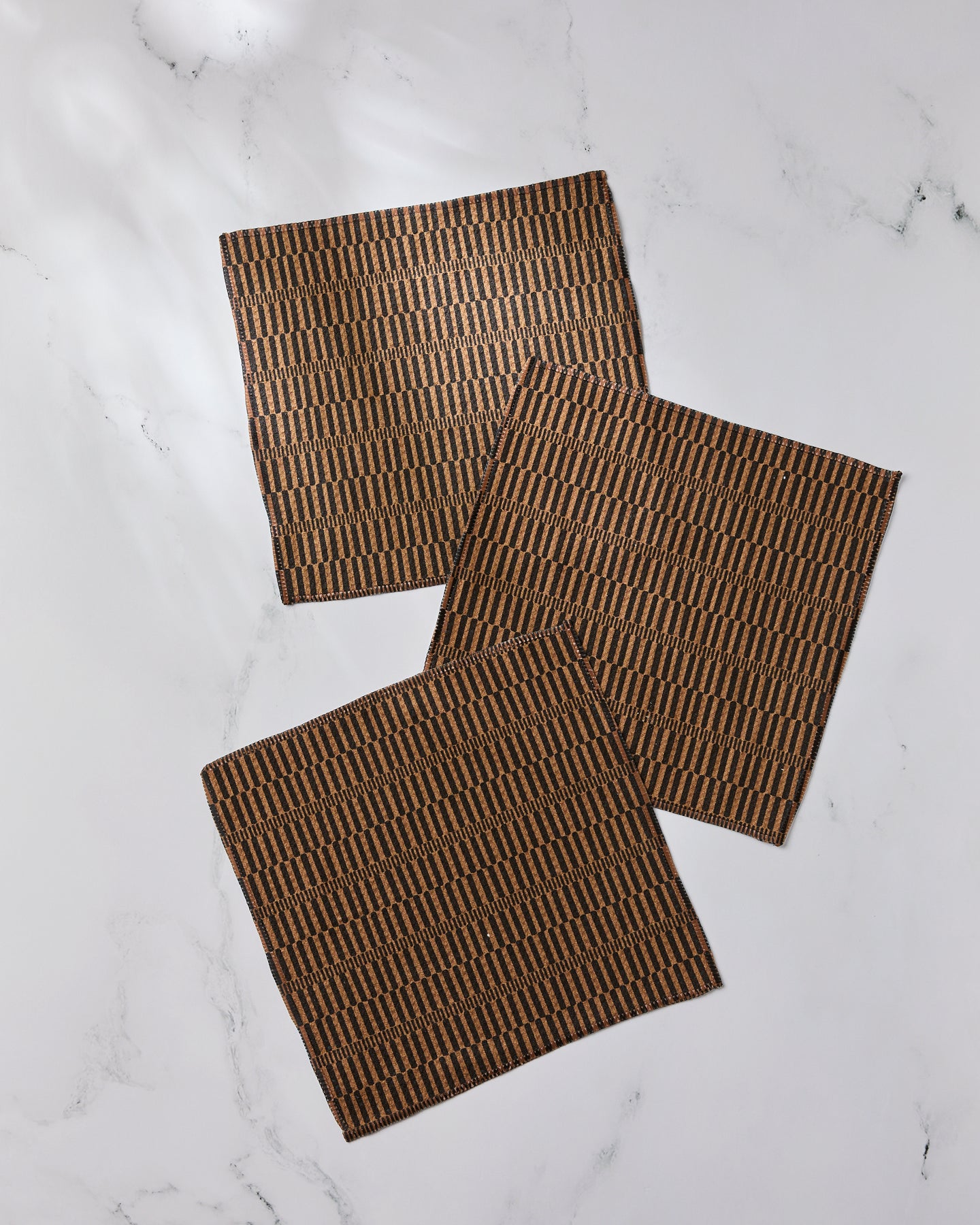 Three Ridge napkins by Geometry, made from recycled materials, feature a black geometric pattern on brown fabric and are arranged on a white marble surface.