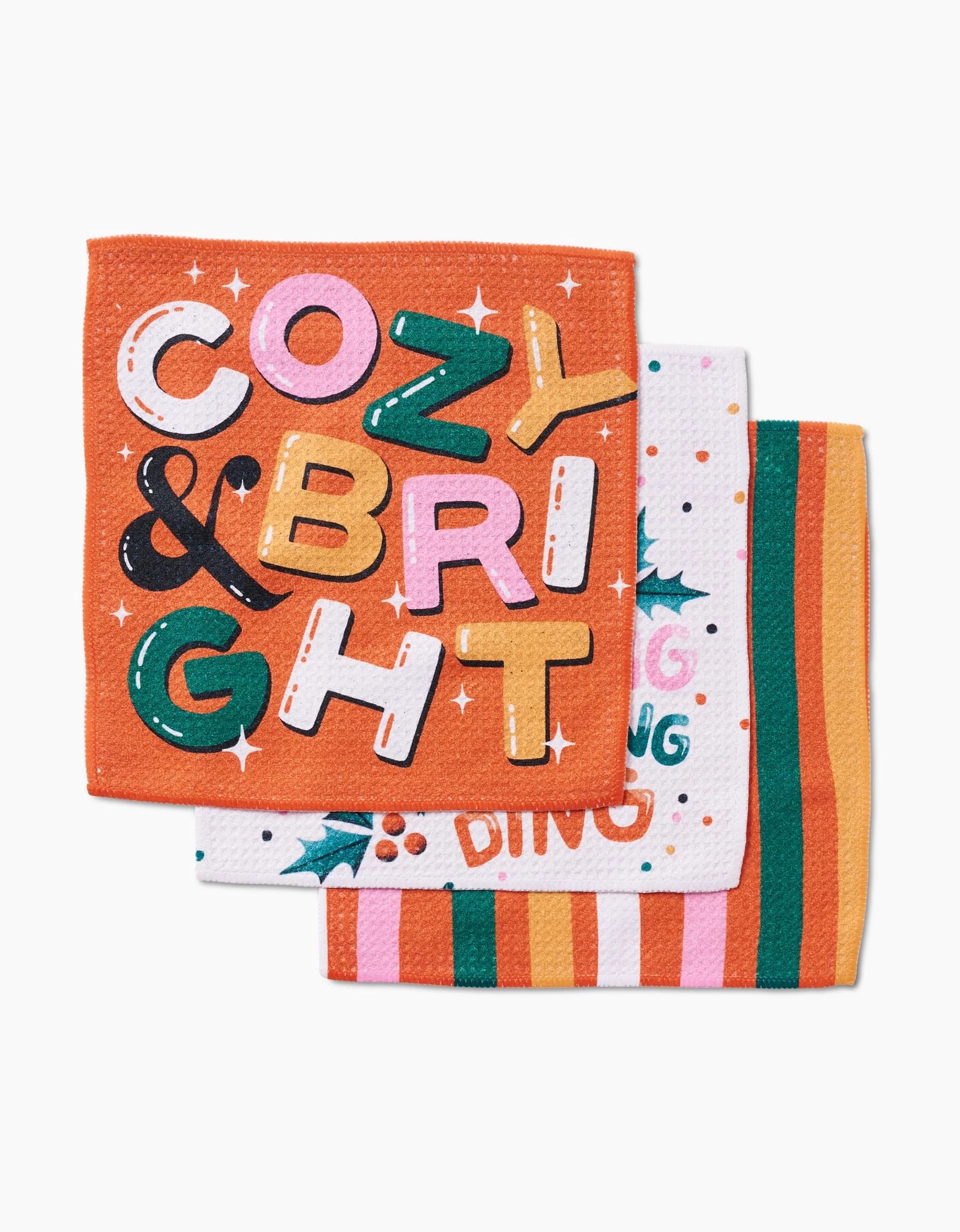 The Megan Roy Ring-A-Ling set features three recycled napkins: the top is orange with COZY & BRIGHT in colorful letters, the middle has multicolored text with holly, and the bottom displays vertical orange, yellow, pink, green, and white stripes.