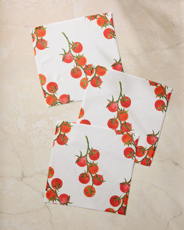 Three "Ripe and Ready" kitchen towels by Geometry, featuring a white base with red tomato and green stem prints, are displayed on a light beige marble surface.