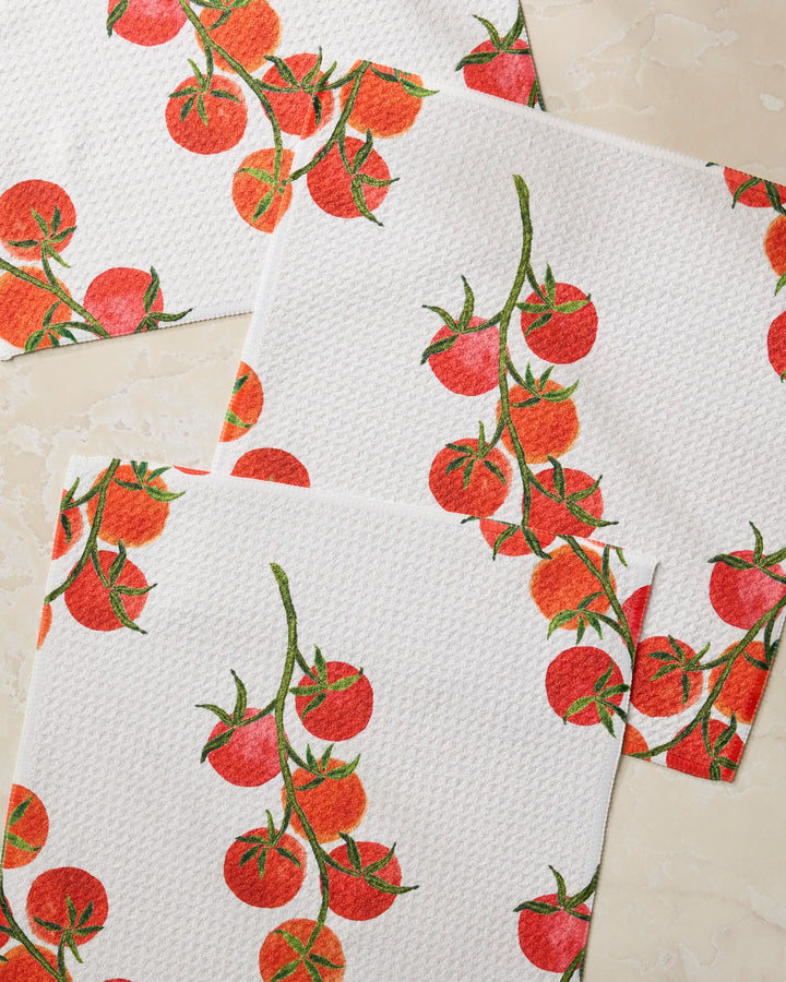 Three white textured napkins from Geometry’s "Ripe and Ready" set display red cherry tomatoes on green vines and are arranged on a light beige marble surface.