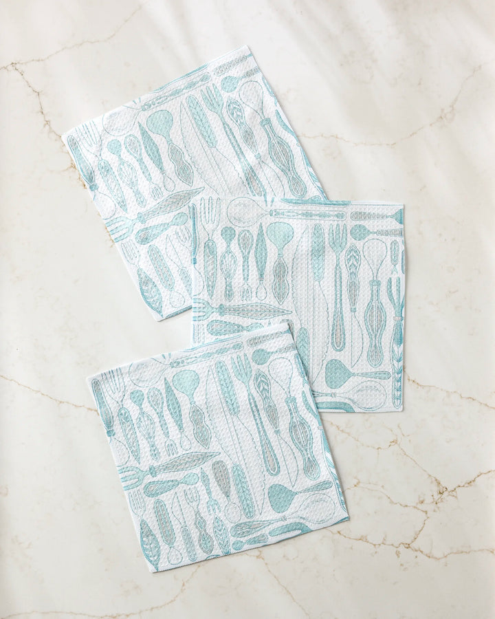 Three Romy Wares blue and white super-absorbent dishtowels by Amy MacCready, featuring various utensil patterns, are arranged with slight overlap on a light marble-like surface.