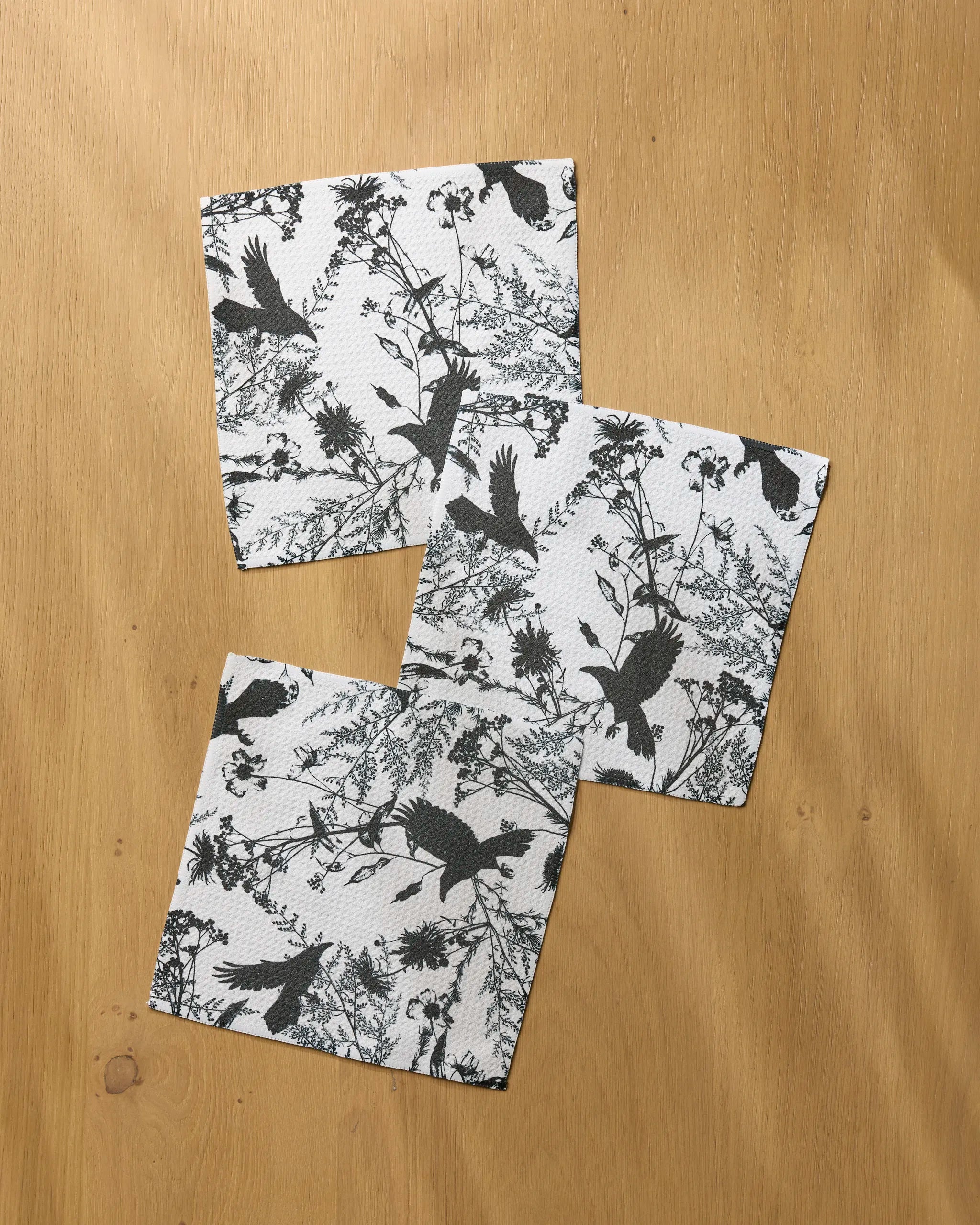 Three Rookery napkins by Geometry, featuring black bird and foliage designs on white recycled fabric, are arranged overlapping on a light wooden surface.