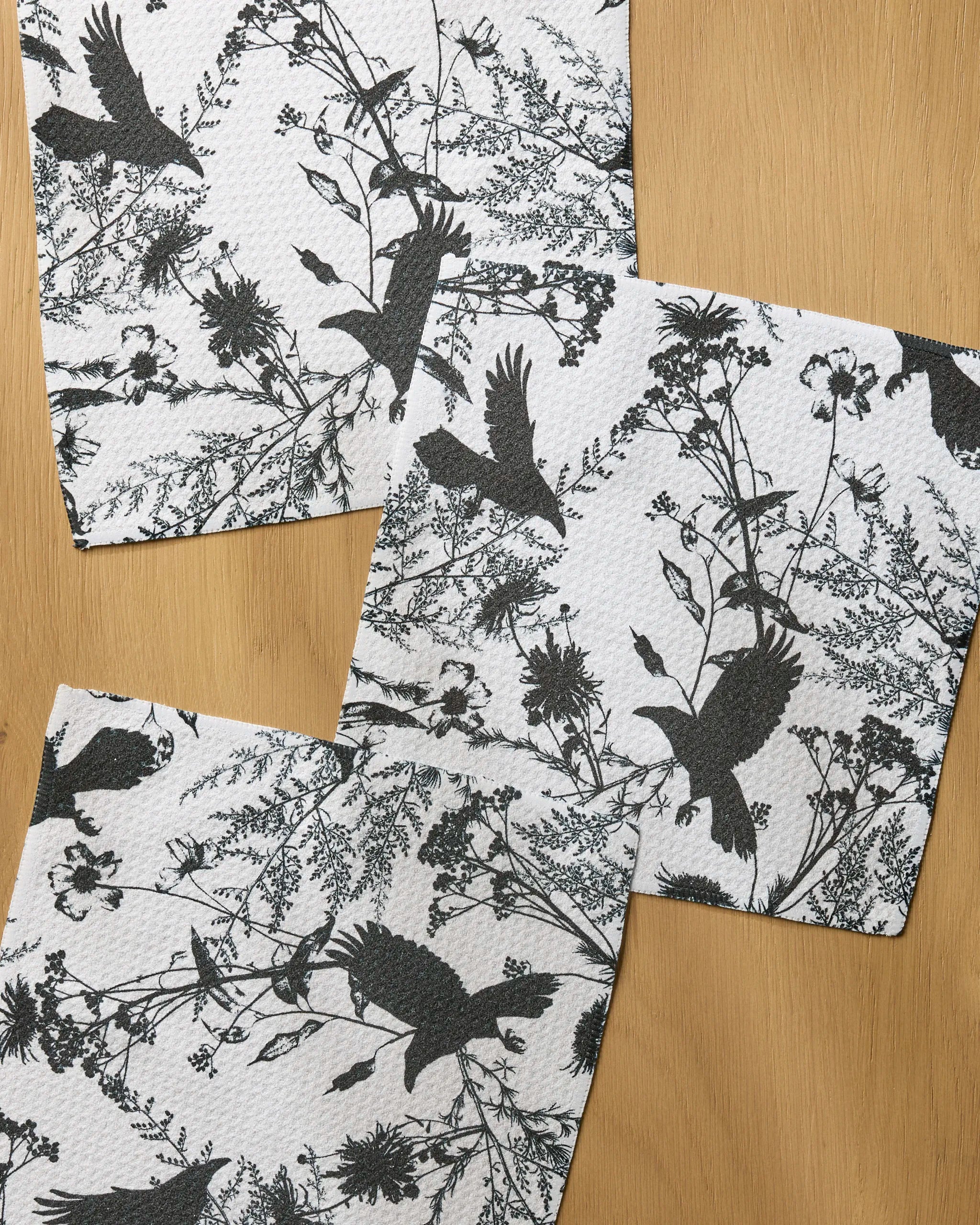 Three white Geometry Rookery dishcloths with black bird and floral designs are arranged on a wooden surface, slightly overlapping.