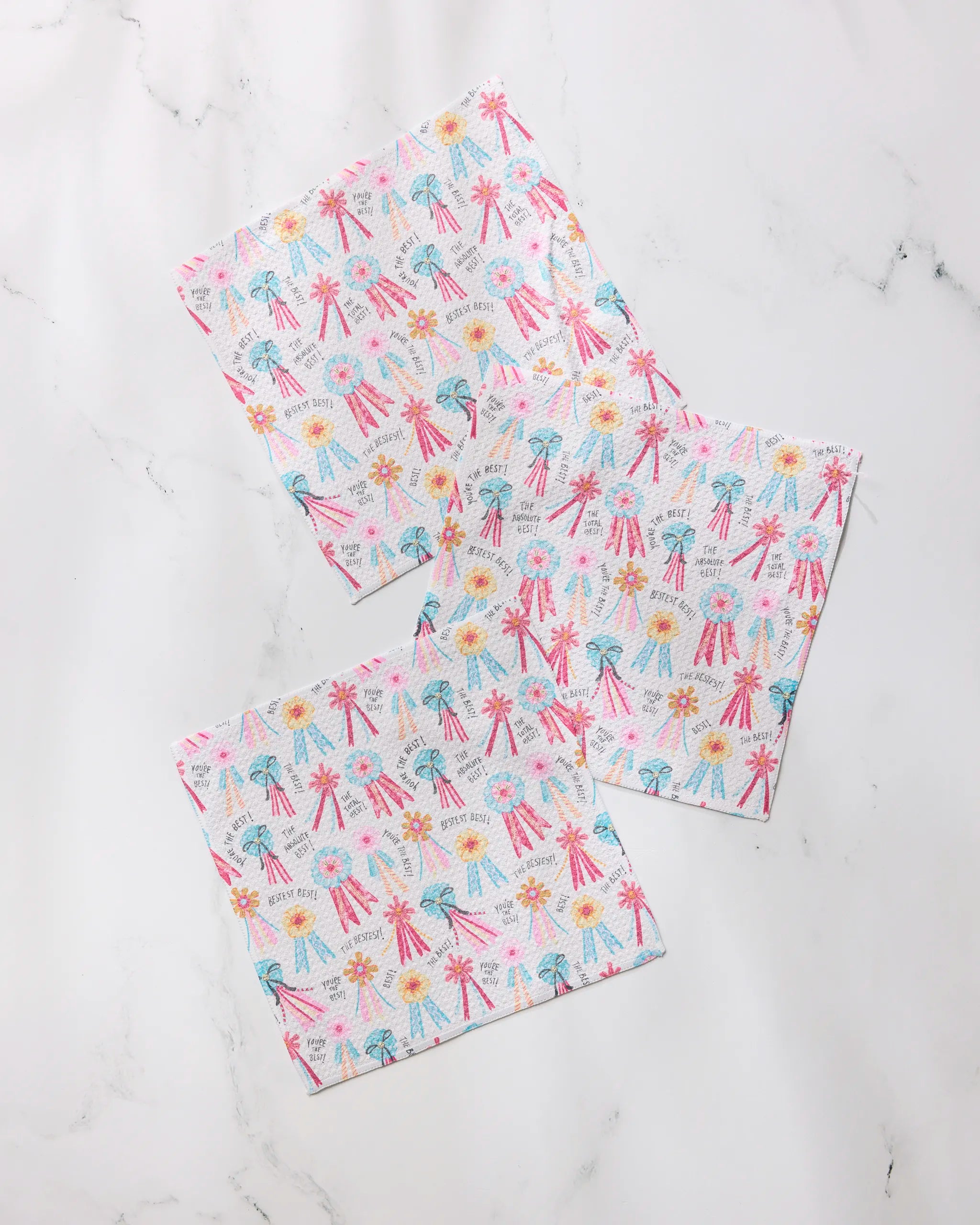 Three Rosettes for You square paper napkins by Geometry, featuring pink, yellow, and blue fireworks and stars, are arranged on white marble—a festive complement to any dishcloth set.