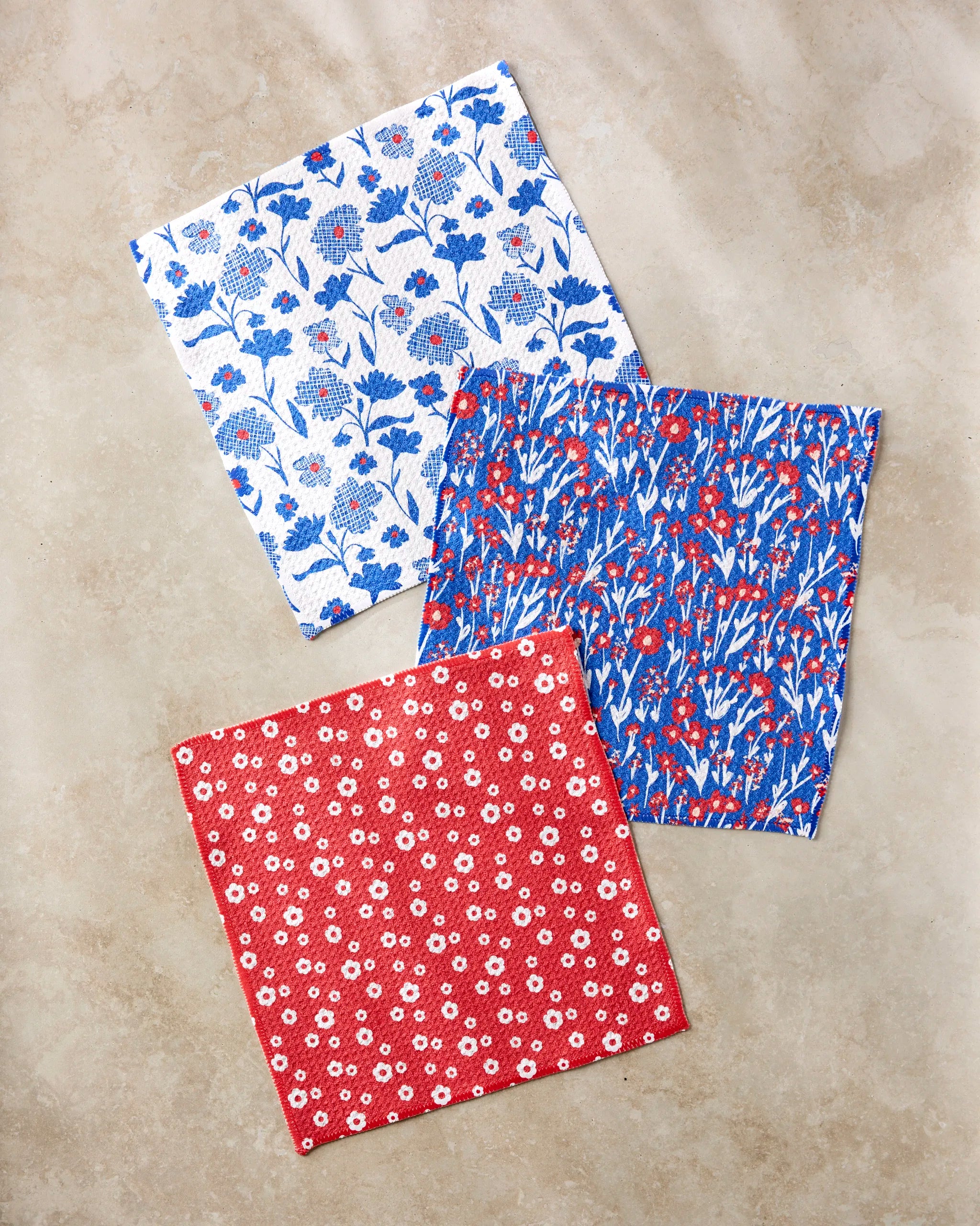 Three Royal Garden cloth napkins by Danelys Sidron, each with a unique floral pattern—white with blue and red flowers, blue with a red and white floral design, and red with small white flowers—are displayed on a beige surface.