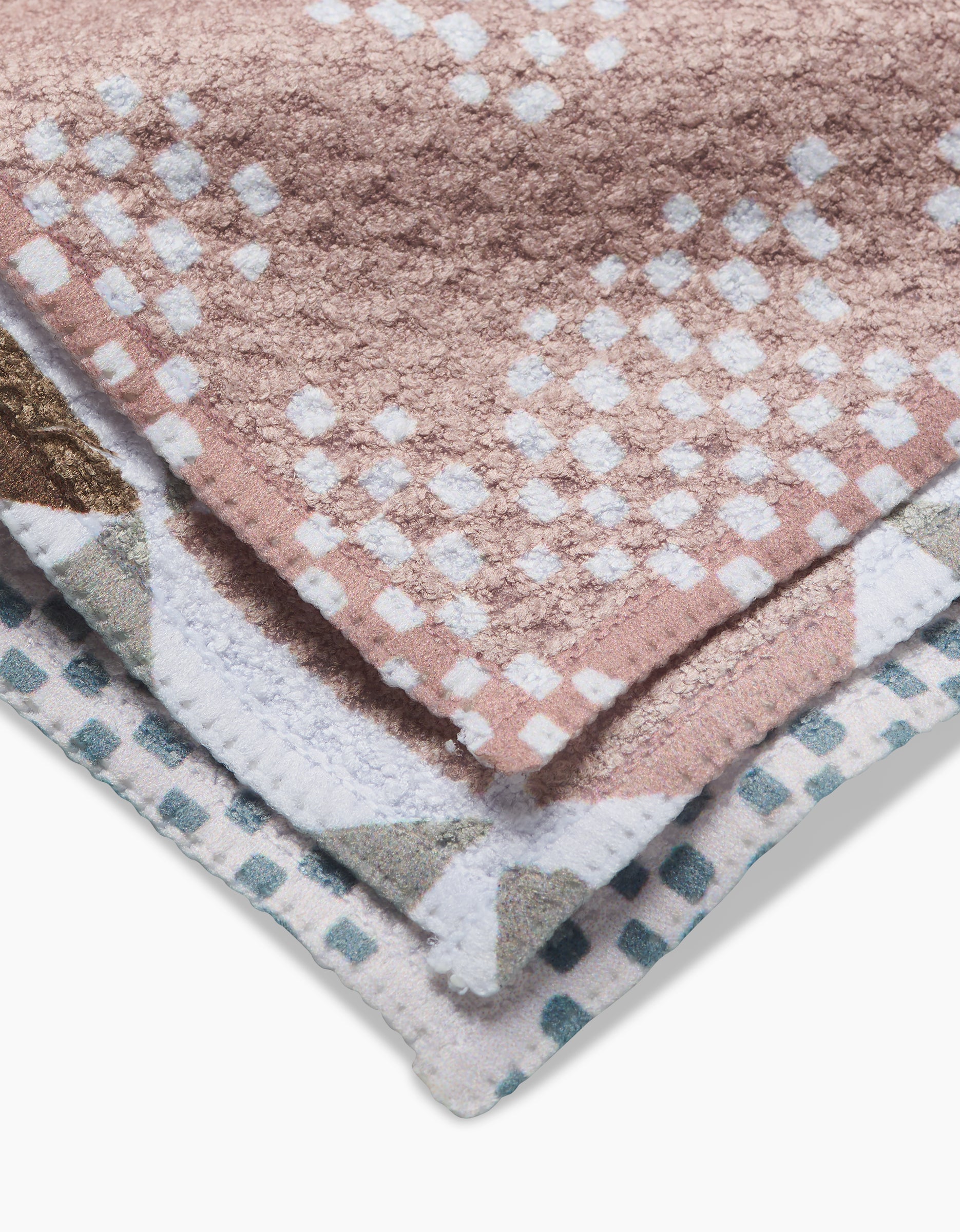 A close-up of two folded Rubix Pixie towels by Studio Jeannot, made from recycled materials—one pink with white and light blue dots, the other white with blue, green, and brown geometric accents.