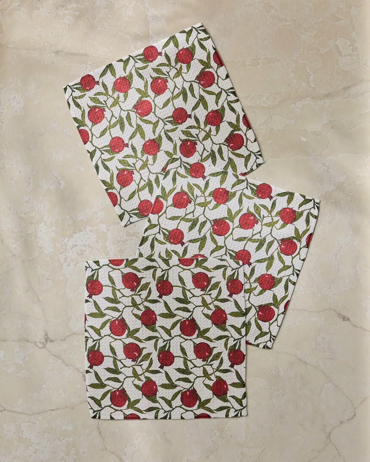 Three Geometry “Ruby Rind” white cloth napkins, featuring a vibrant red pomegranate and green leaf pattern, are displayed on a beige marble surface—bringing a charming accent to your table or serving as an eco-friendly upgrade to your collection of recycled dishcloths.