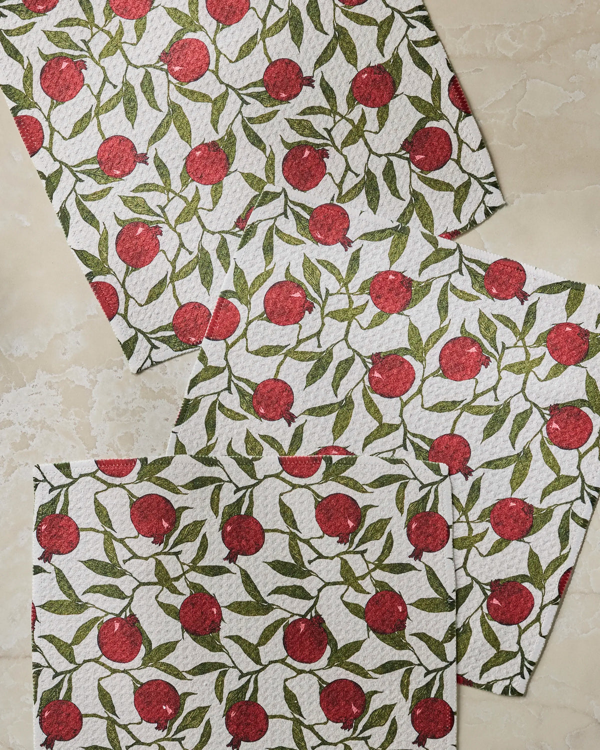 Three textured, absorbent Geometry kitchen towels in the Ruby Rind design—featuring vibrant red pomegranates and green leaves—are arranged diagonally and overlapping on a beige marble surface.