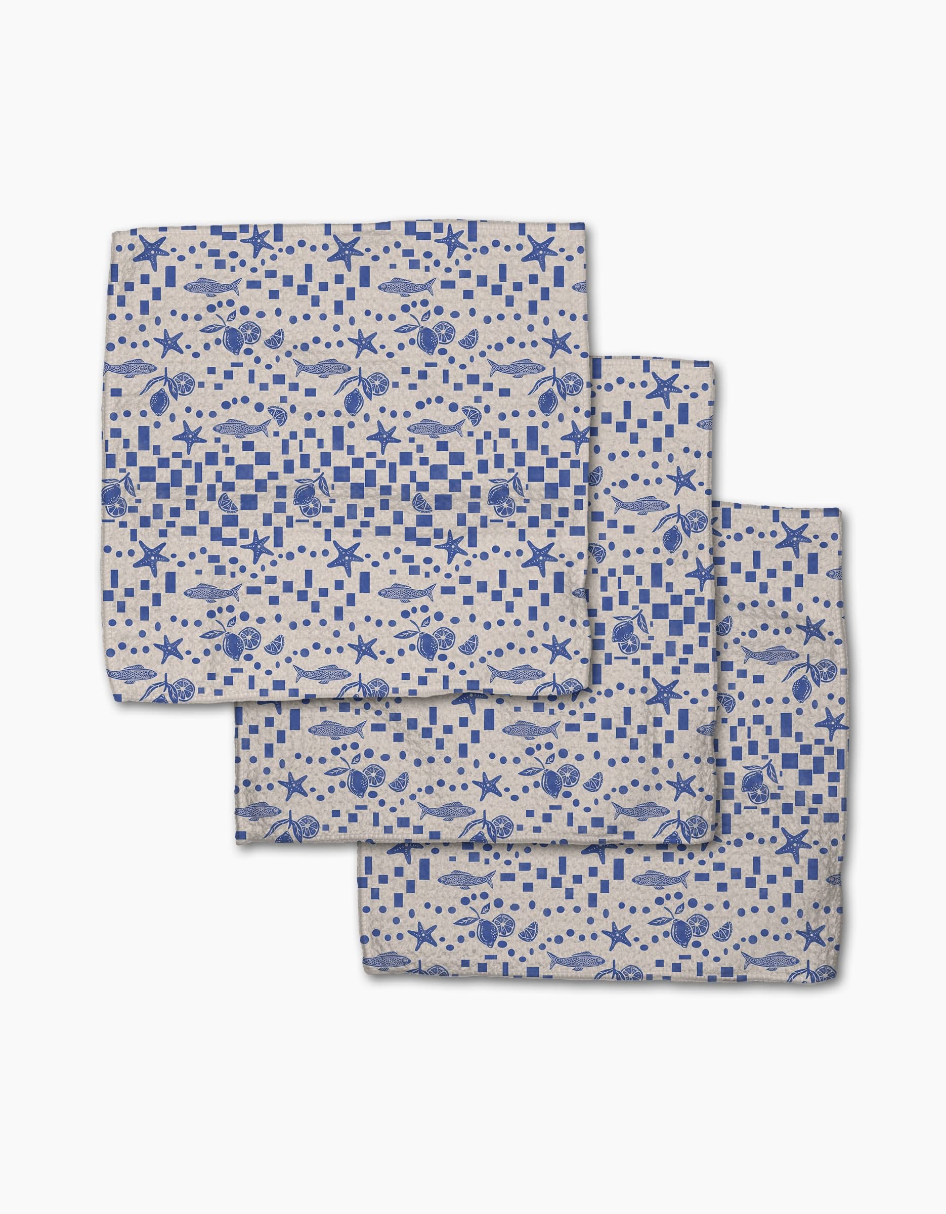 Three folded Geometry Santorini Tile dishcloths, featuring blue nautical patterns of fish, crabs, seashells, and checkered squares on beige Geoweave fabric, are arranged overlapping on a white background.