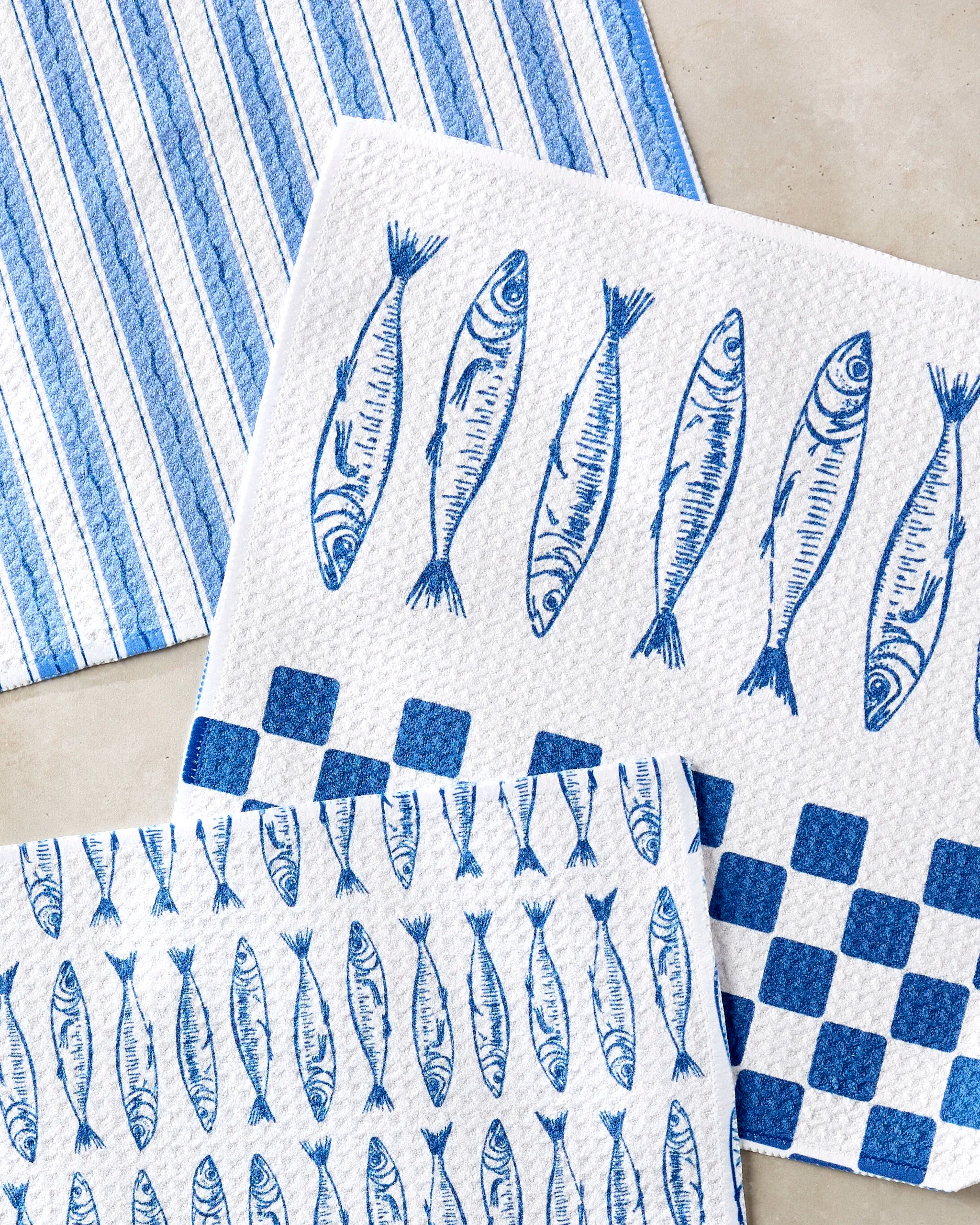 The Sardine Check Blue dishcloth set by Hufton Studio includes three blue and white super-absorbent dishtowels with unique patterns: stripes, large fish with checkered squares, and repeating small fish, displayed on a light surface.