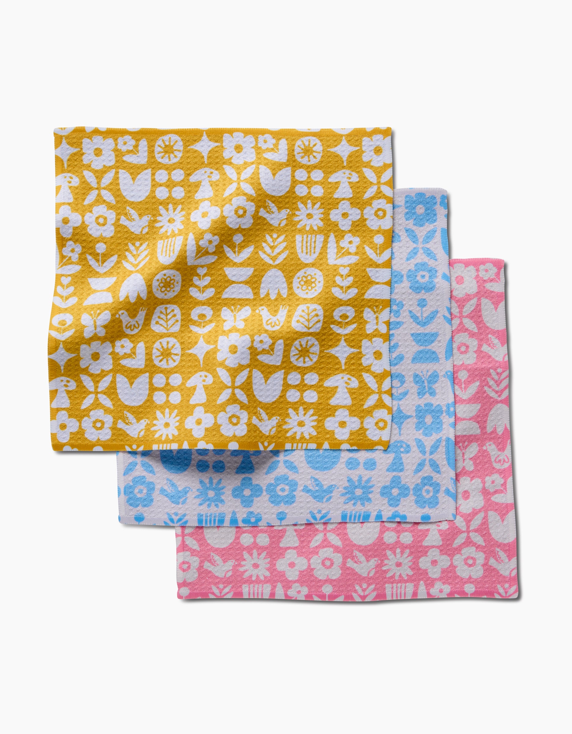 Three Kayla Ann Scandi Spring dishcloths—yellow, blue, and pink—feature white floral and botanical prints. These super-absorbent Geoweave cloths are neatly overlapped on a white background.