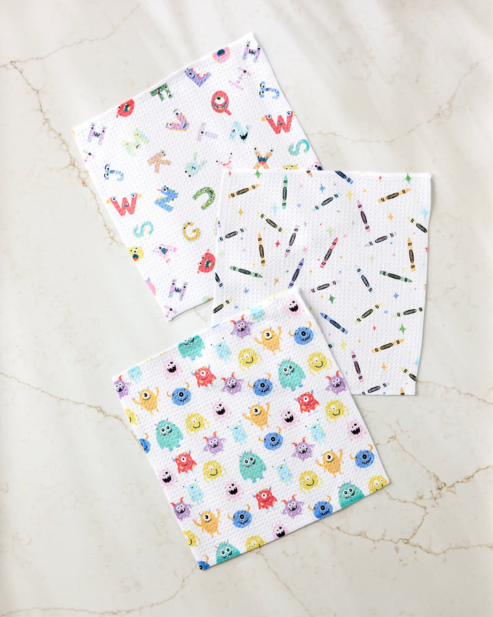 Three Scribble Squad absorbent kitchen towels by Vaishali Design Studio are displayed on a light marble surface: one with playful letters and animals, another with scattered crayons, and the third featuring cheerful cartoon monsters.