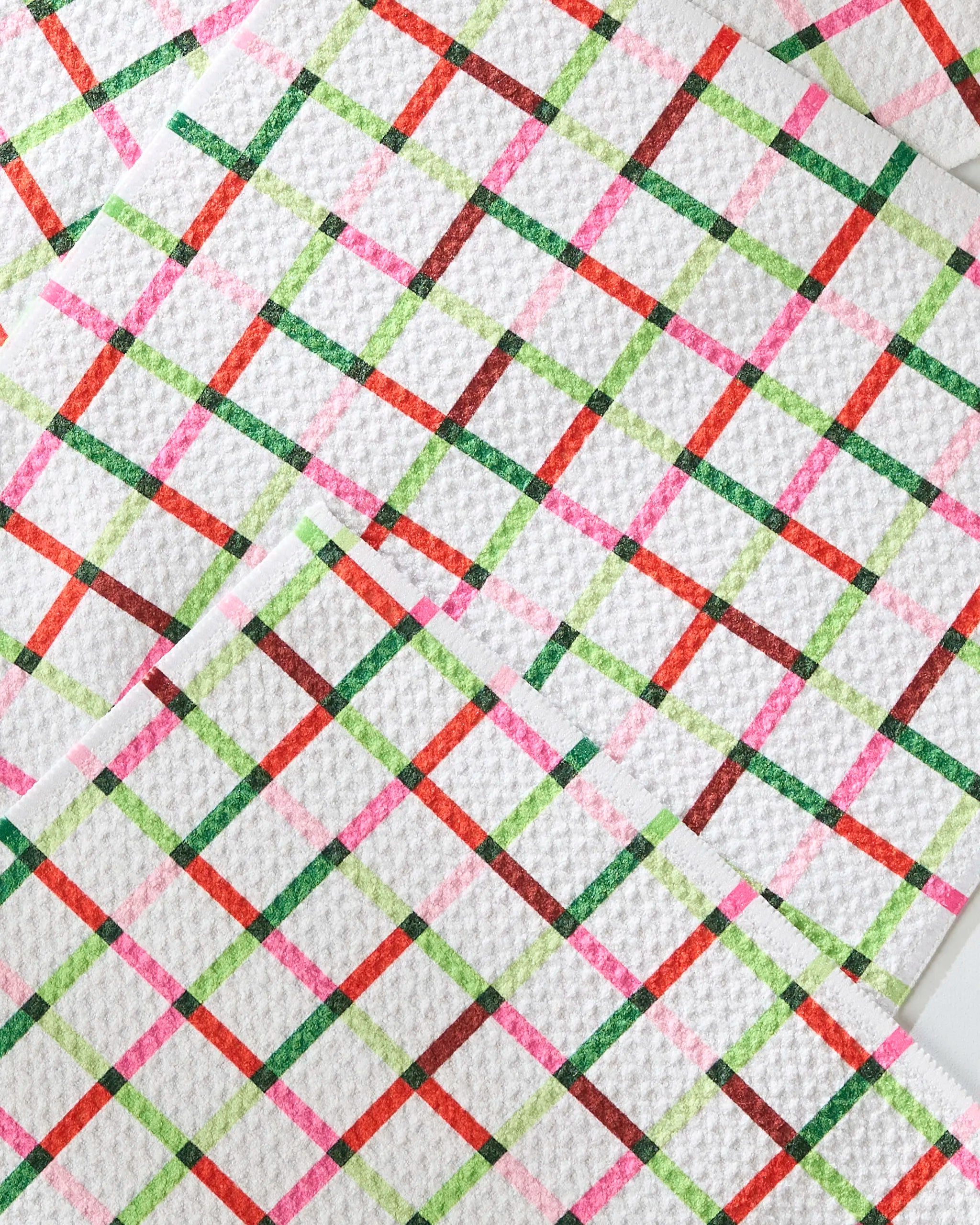 A close-up of Geometry’s Season of Joy paper towels shows textured white recycled material adorned with a vibrant diagonal crisscross pattern in red, pink, green, and brown lines.