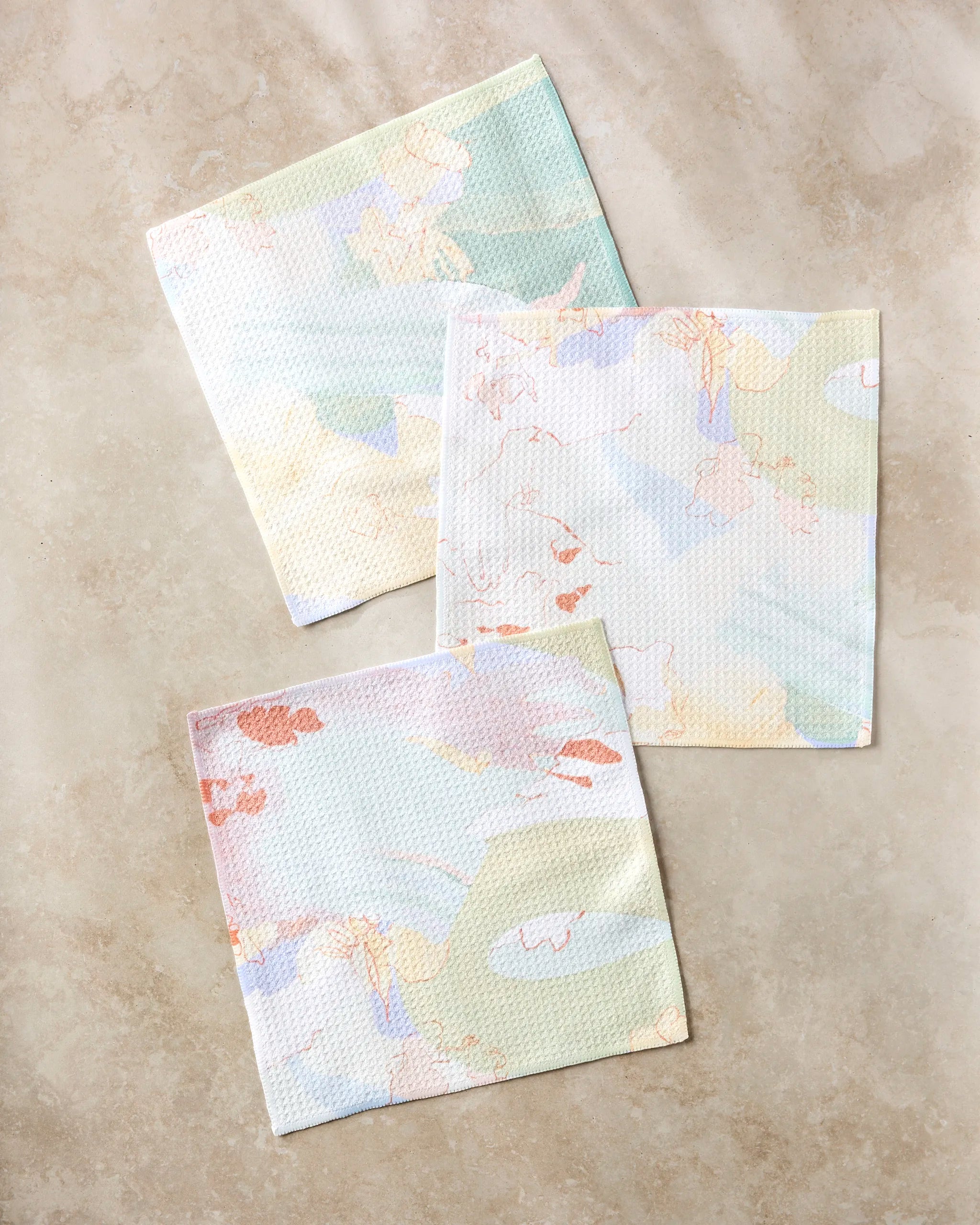 The Secret Fort dishcloth set by Kristina Baker features three pastel reusable cloths made from recycled materials in soft green, yellow, peach, and blue shades with abstract patterns, arranged on a light beige surface.