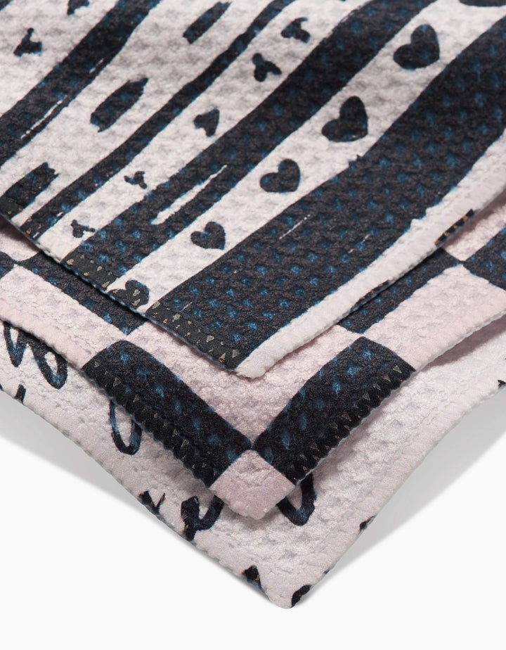 A close-up of Danelys Sidrons Self Love dishcloths shows three folded Geoweave towels with black stripes, hearts, and dots on white and pale pink, featuring visible stitching for added texture.