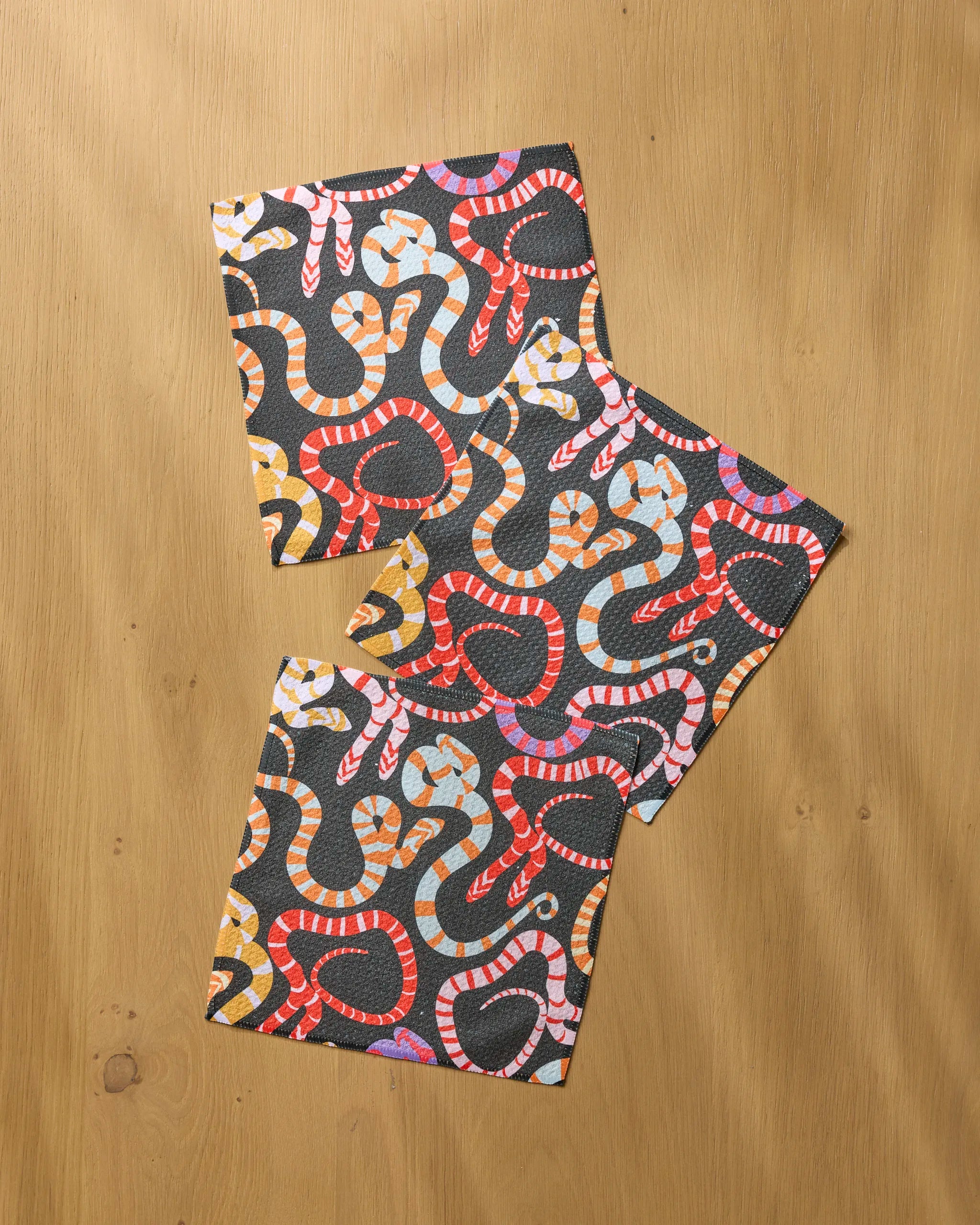 Three absorbent Serpentine Riot dishcloths by Geometry, each featuring colorful snake patterns in red, pink, orange, and yellow on dark backgrounds, are displayed diagonally on a light wooden surface.
