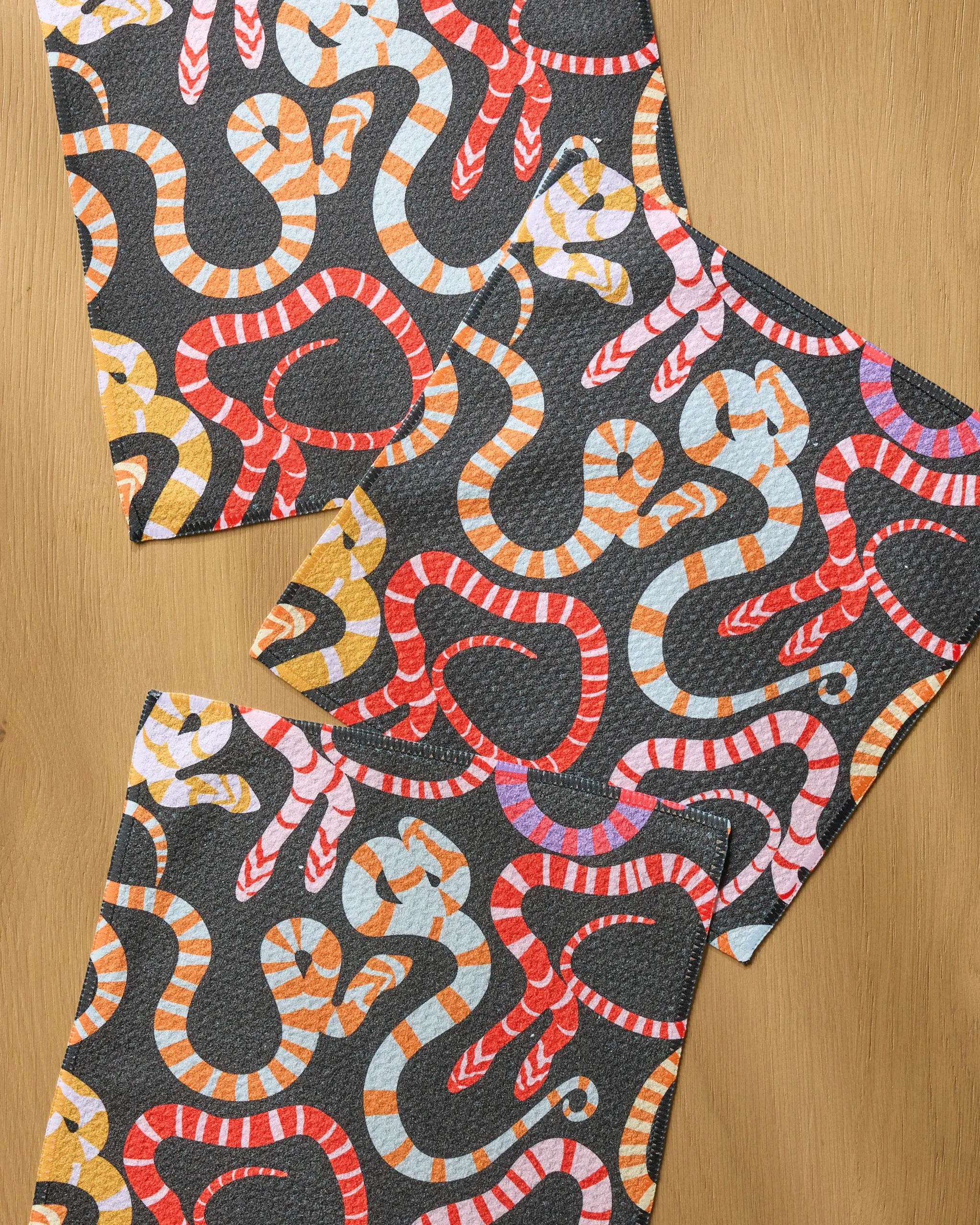 Three Geometry Serpentine Riot dishcloths featuring colorful red, orange, yellow, and white striped snake patterns on a dark background are displayed on a wooden surface.
