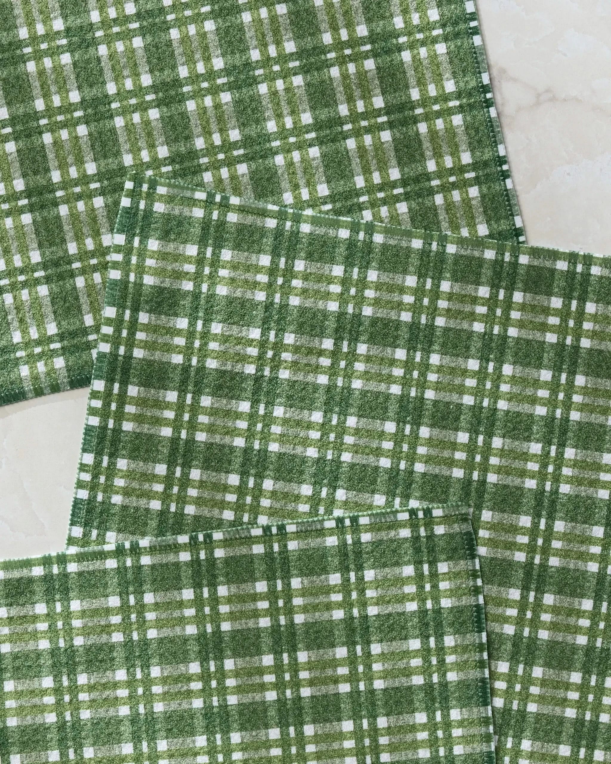 Four Shay Plaid cloth napkins by Geometry, made from recycled materials, are arranged overlapping on a light marble-style surface. The green plaid pattern features various shades of green with white stripes.