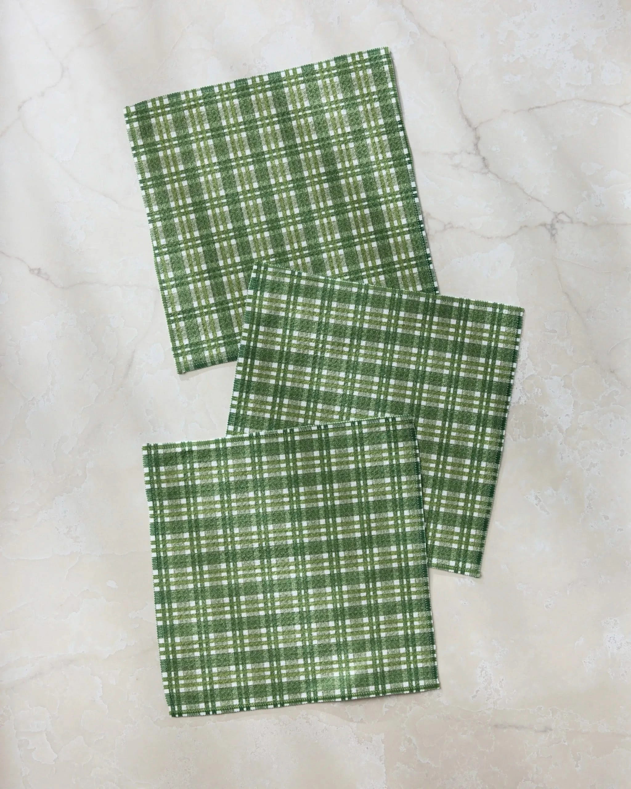Three Shay Plaid napkins by Geometry, from a dishcloth set, are displayed on a light marble-like surface, each green and white napkin slightly overlapping the others.