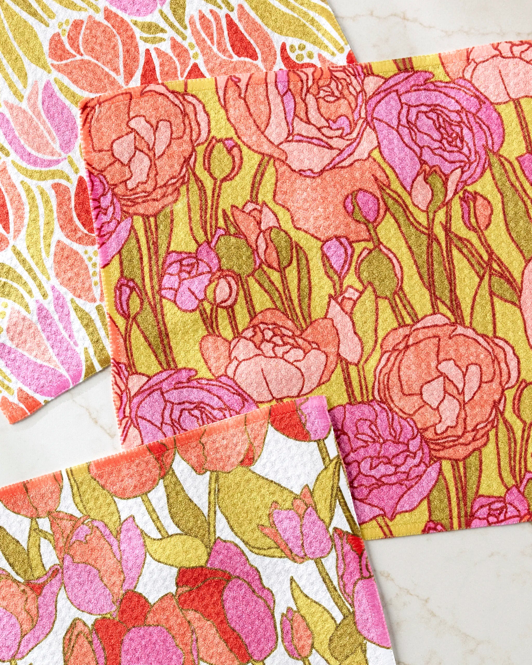 The Sidney dishcloth set by Amy MacCready features three rectangular cloths with vibrant floral and leaf patterns in pink, red, orange, and yellow. Made from recycled materials, they’re displayed on a light marble surface.