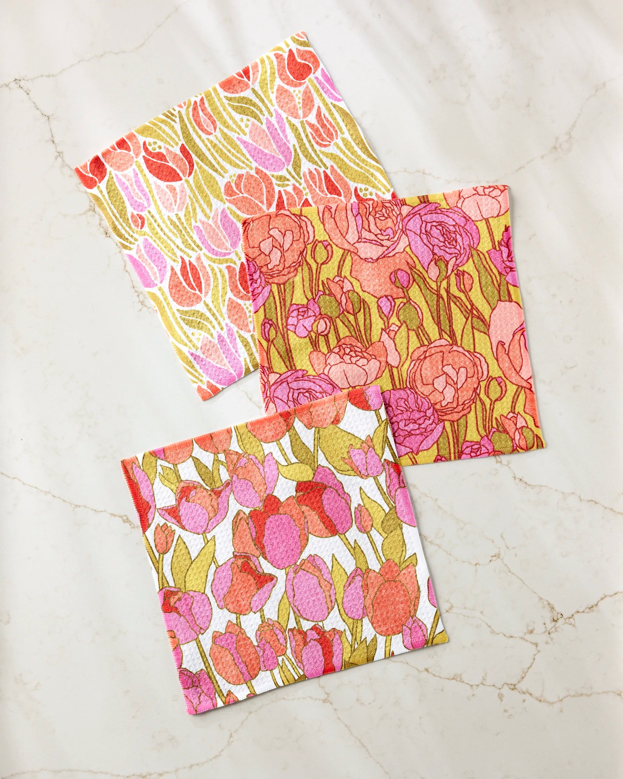 Three square cloths from the Amy MacCready Sidney set, made of recycled materials, feature pink, red, and orange floral patterns on assorted backgrounds, shown overlapping on a white marble surface.