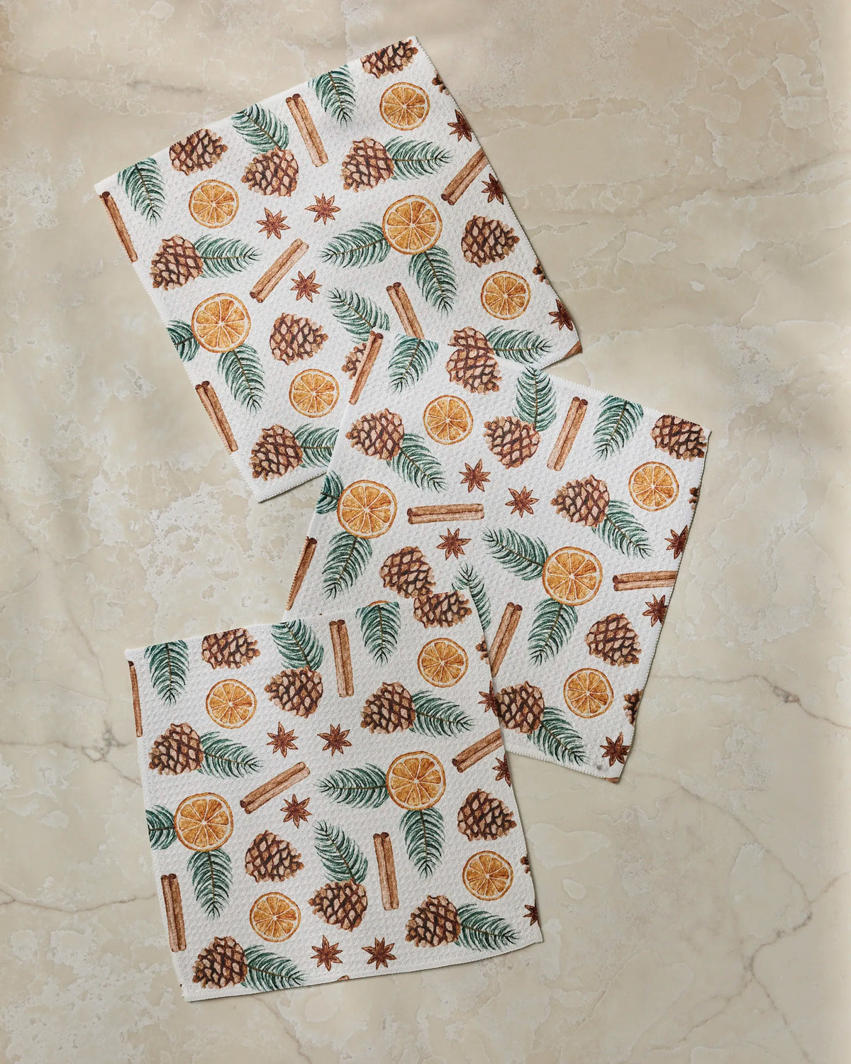 Three patterned absorbent dishcloths from Geometry’s Simmer Pot collection, featuring designs of pinecones, orange slices, cinnamon sticks, star anise, and pine branches, are arranged on a beige marble surface.
