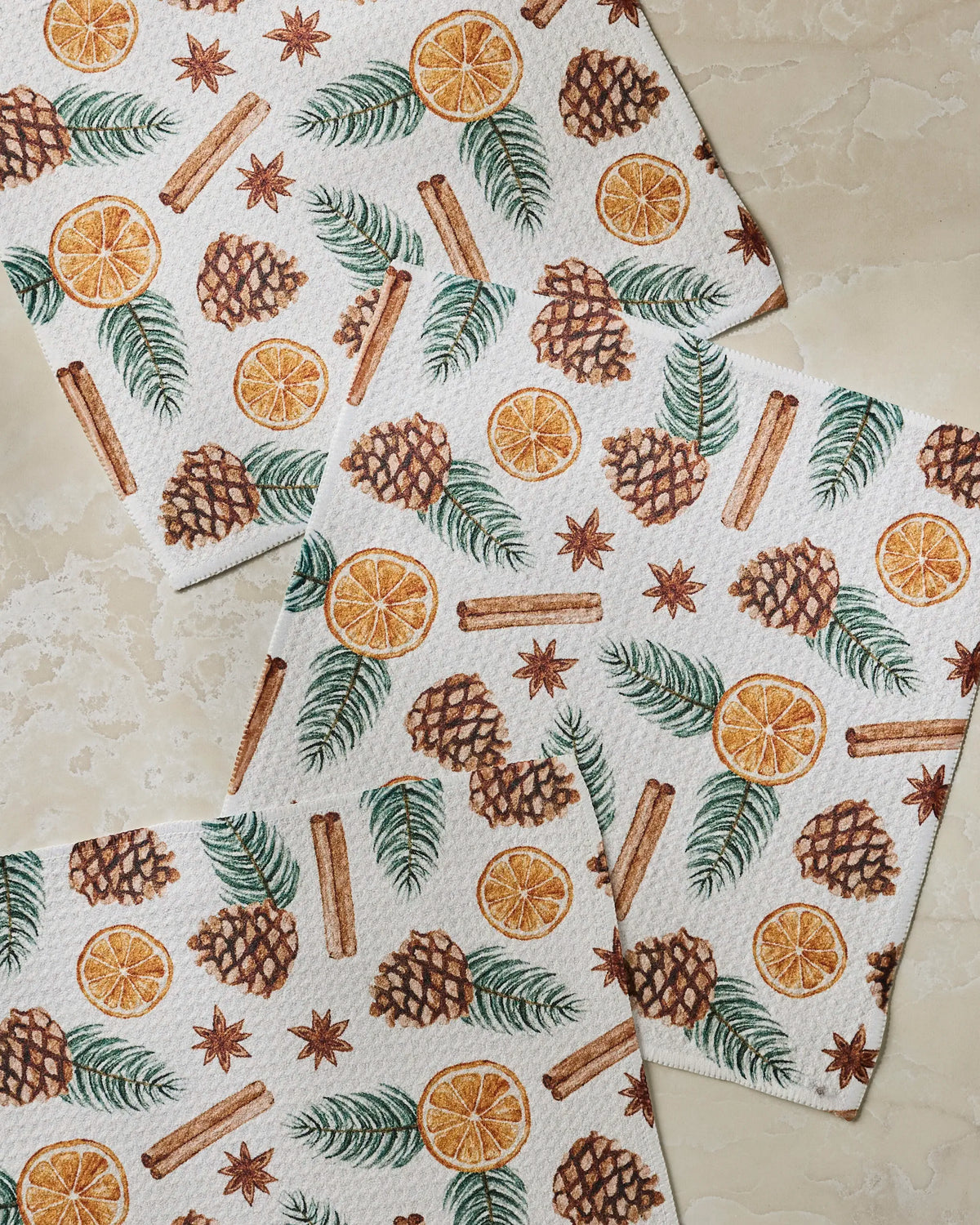 Three Simmer Pot paper towels from Geometry, featuring a pattern of pinecones, cinnamon sticks, dried orange slices, anise stars, and pine branches, overlap on a light marble surface.