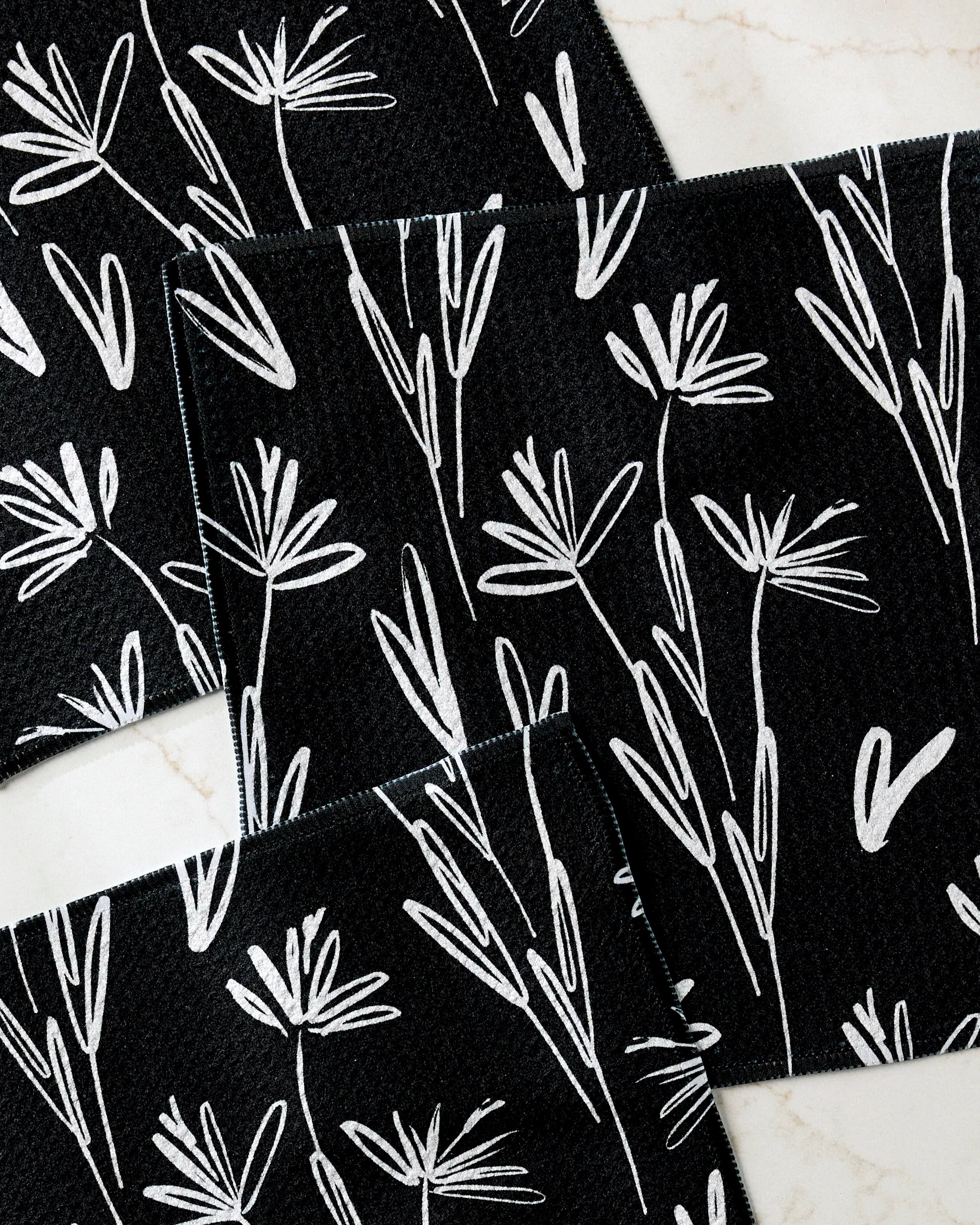 Four black Sketchbook Dusk placemats by Toboggan Avenue, made from recycled materials and featuring white floral line designs, are arranged on a light marble surface with their corners slightly overlapping.
