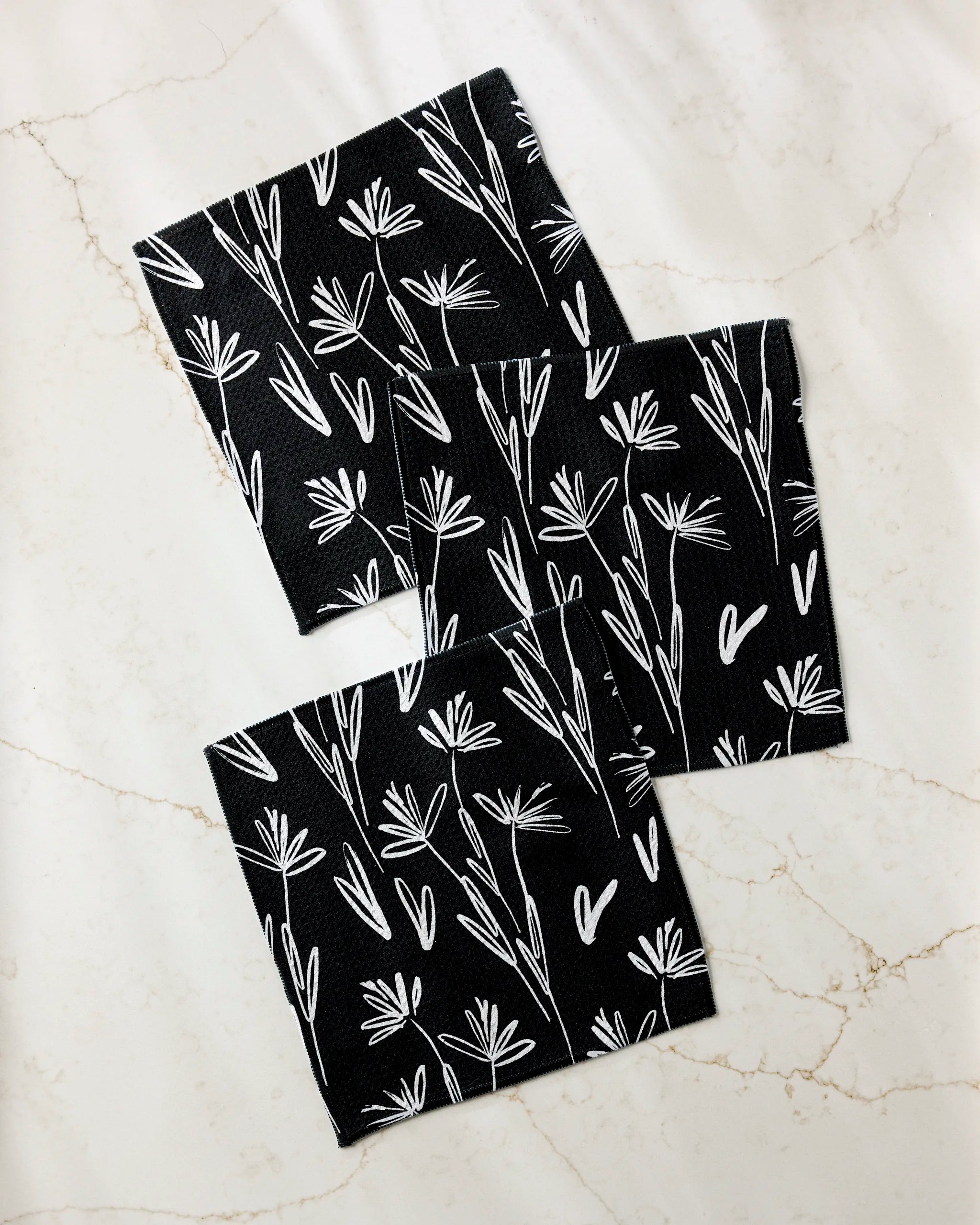Three black square “Sketchbook Dusk” coasters by Toboggan Avenue, featuring white floral line art and made from recycled materials, are displayed on a light marble surface.