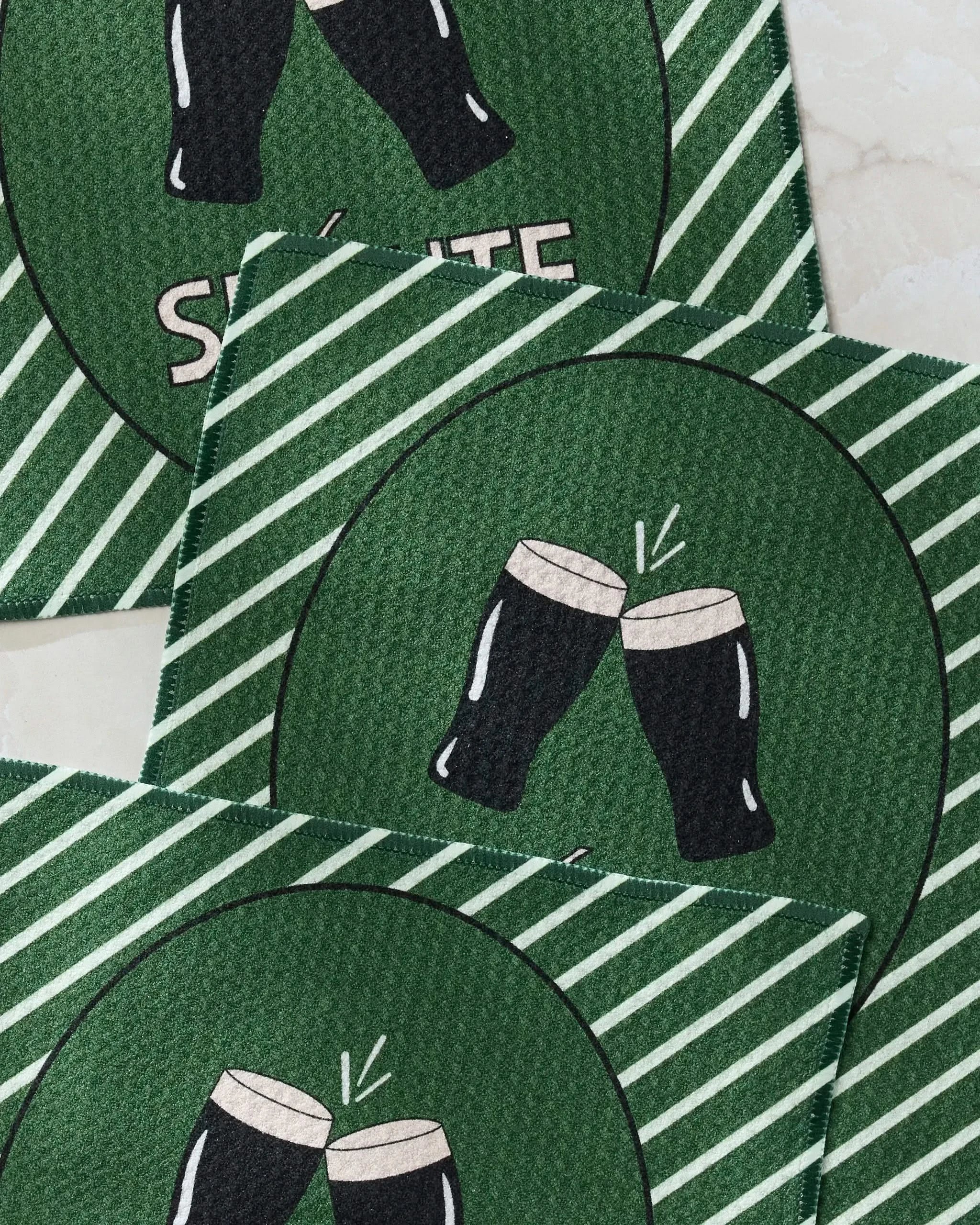 Three green striped "Slainte" drink coasters by Geometry, made from recycled materials, feature two clinking dark beer glasses and partial white "SALUTE" lettering on a light background.