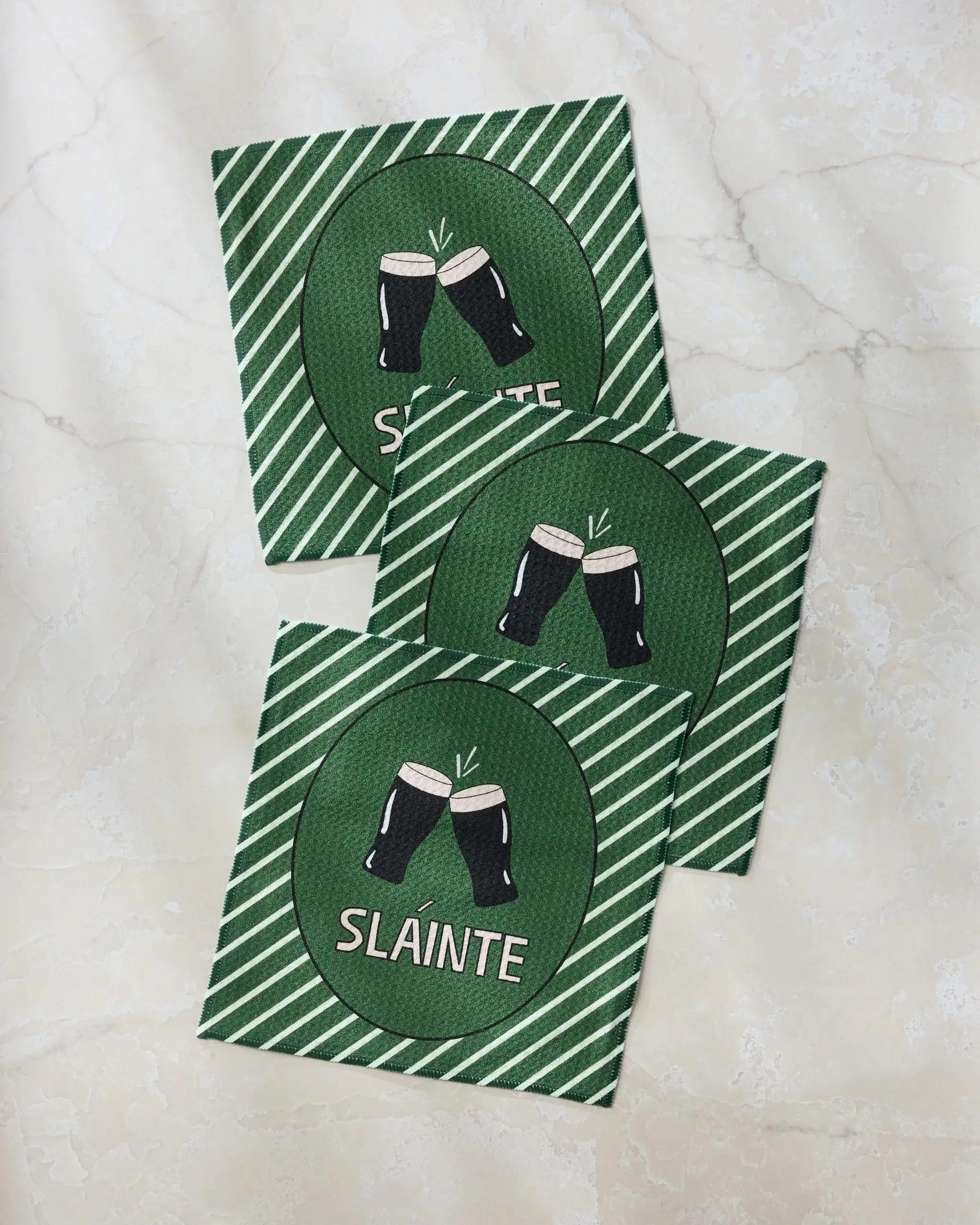 Three green Geometry Slainte napkins with white diagonal stripes and two clinking beer glasses above "Slainte" are arranged on a marble surface—perfect to pair with your favorite recycled kitchen towels.