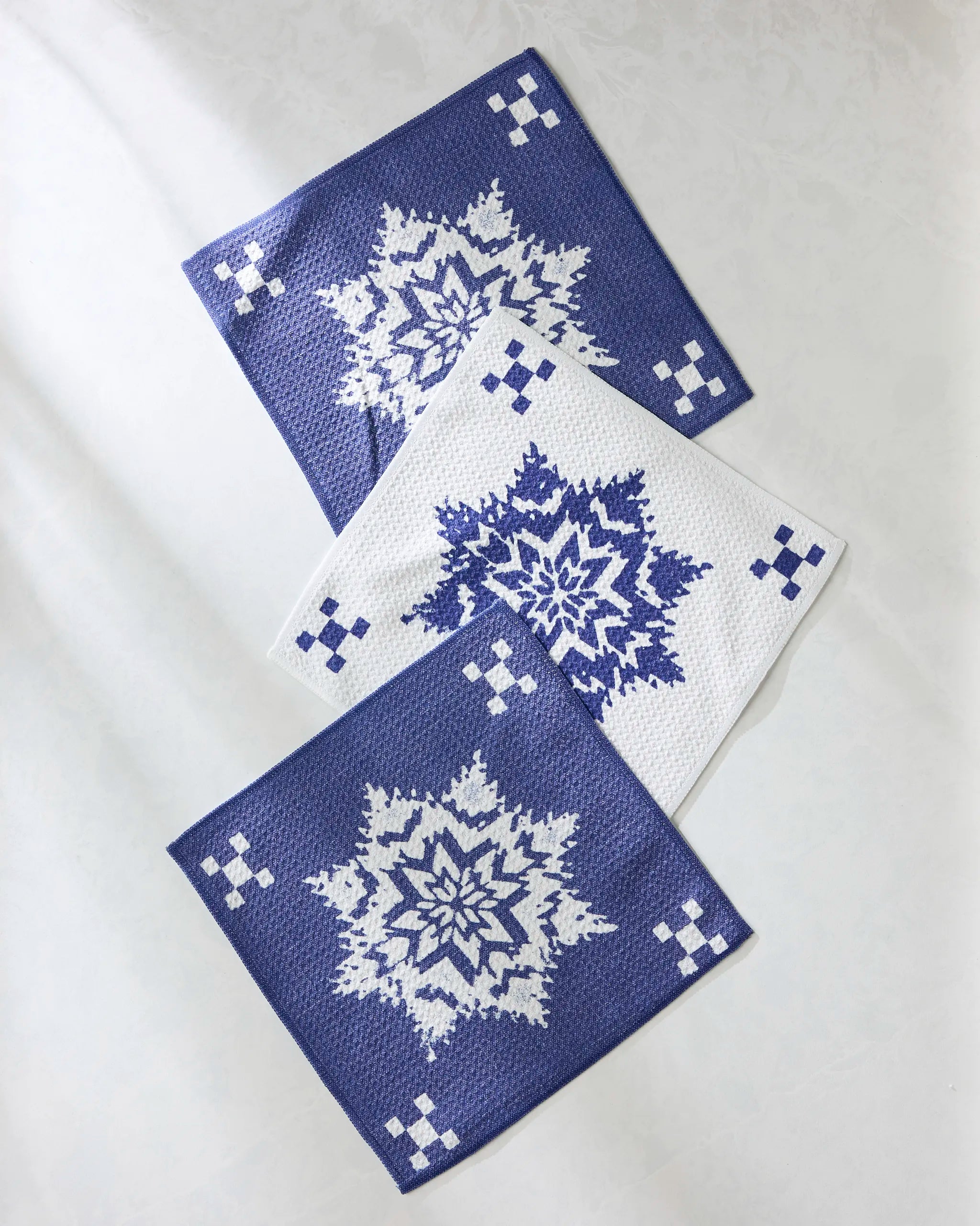 The Geometry “Snowfall” set features three super-absorbent dishcloths: two dark blue cloths with white snowflakes and one white cloth with a blue snowflake design, all arranged on a white surface.