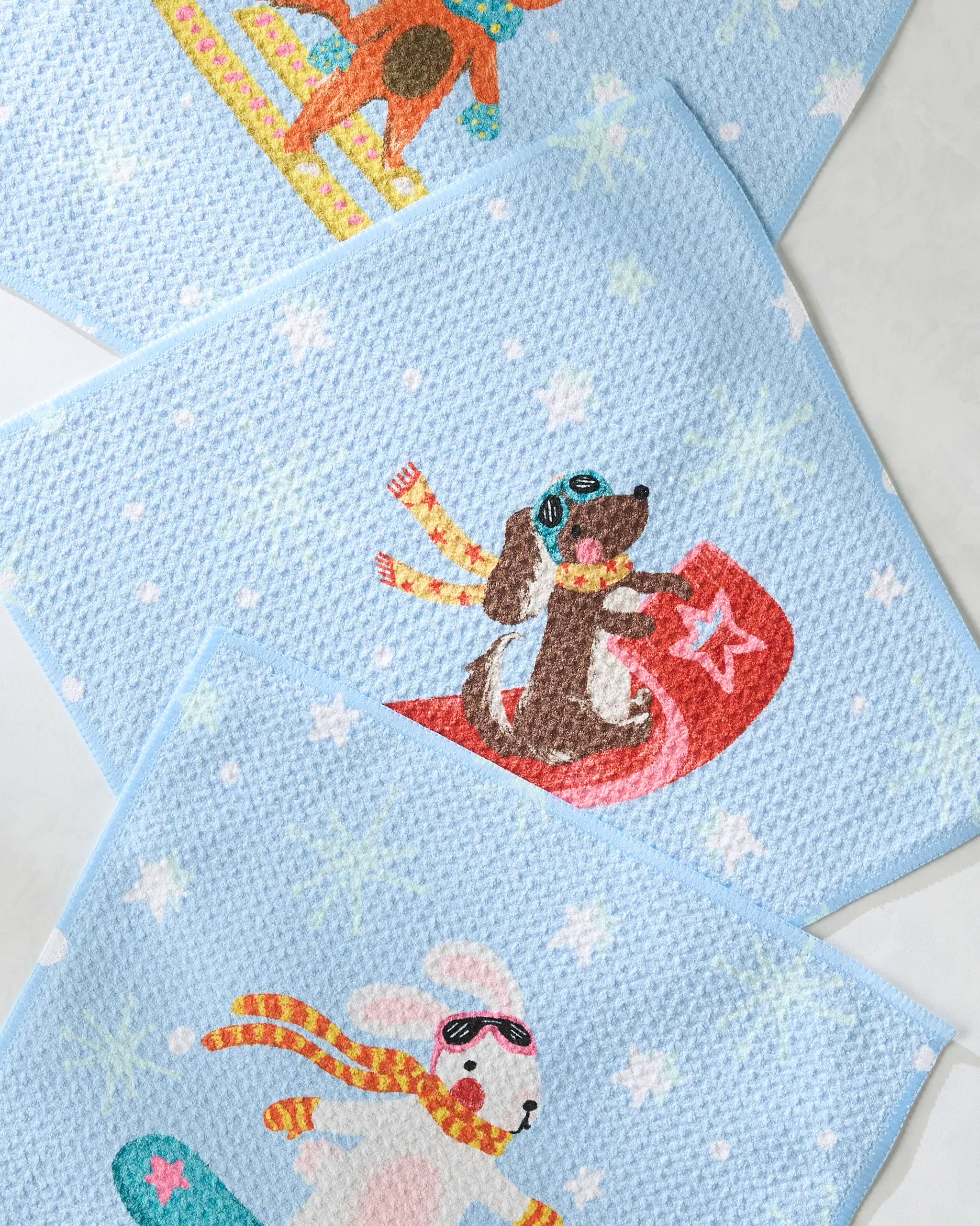 The Geometry Snowy Friends Dishcloth Set includes three blue cloths with playful winter scenes—a dog sledding, a dog on a red sled, and a rabbit snowboarding—made from recycled materials for an eco-friendly, super-absorbent clean.