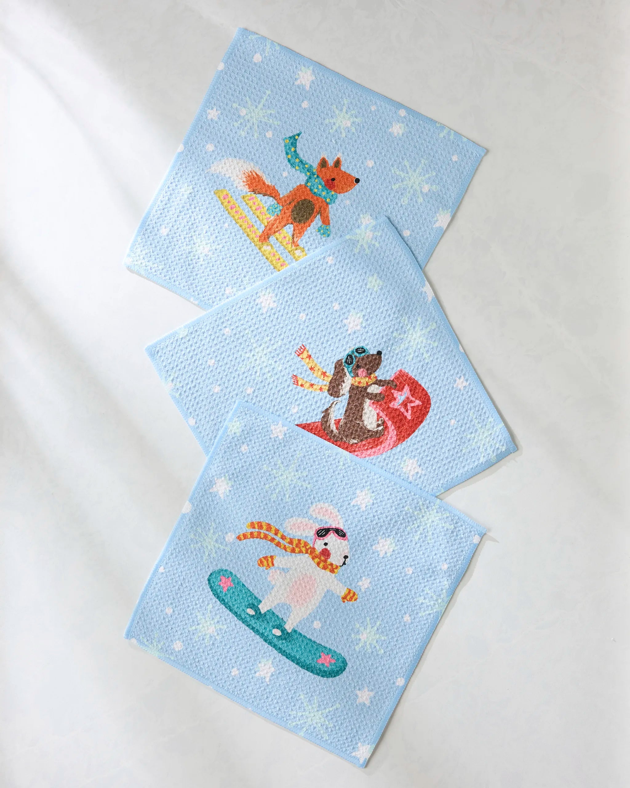 Geometry’s Snowy Friends features three blue, recycled napkins with white stars and snowflakes, each showing a fun cartoon animal snowboarding or sledding—an eco-friendly, super-absorbent addition to your Dishcloth Set.