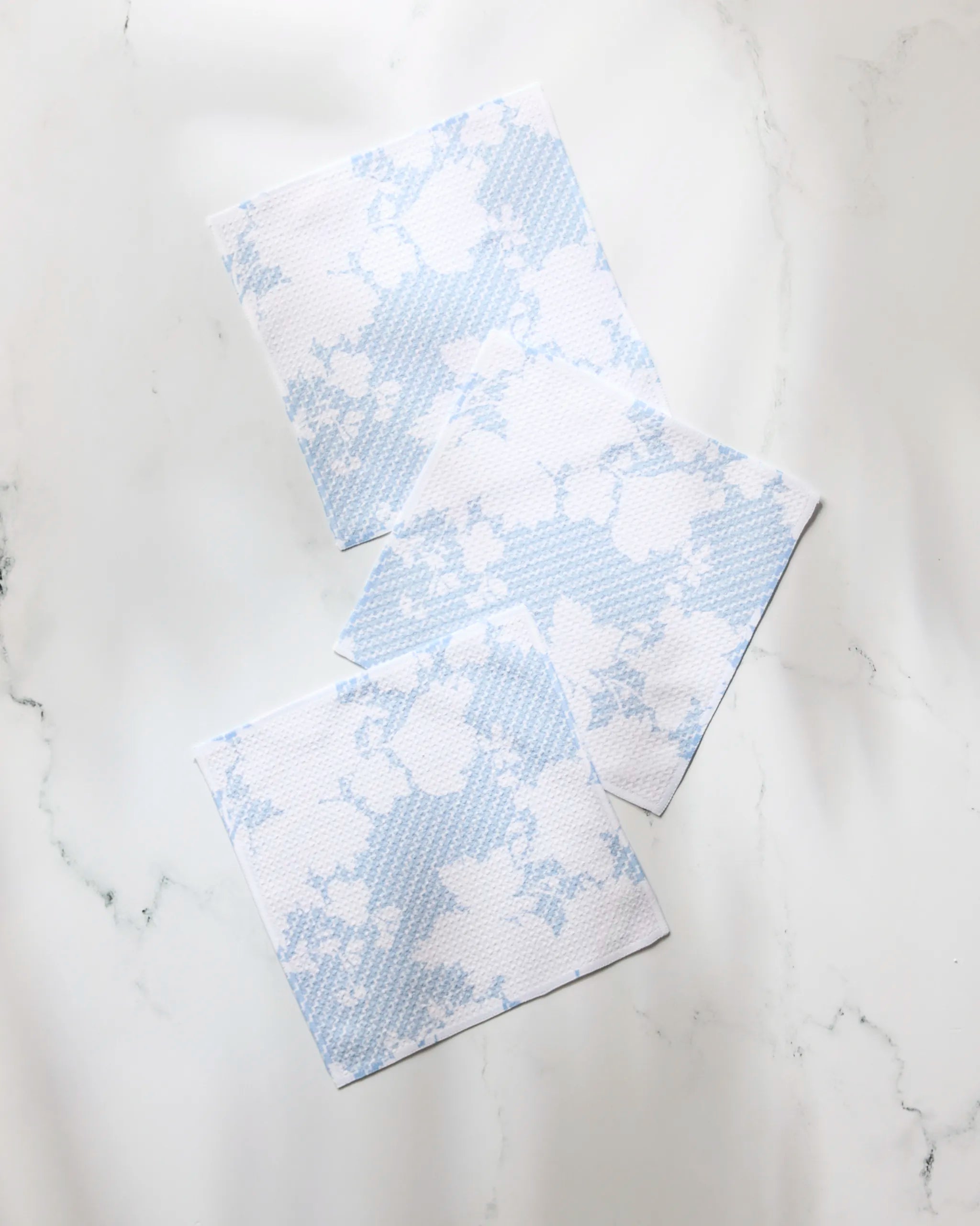 Three white Soft Bloom dishtowels by Geometry, featuring a light blue pattern and made from recycled materials, are arranged in an overlapping, scattered style on a white marble surface.