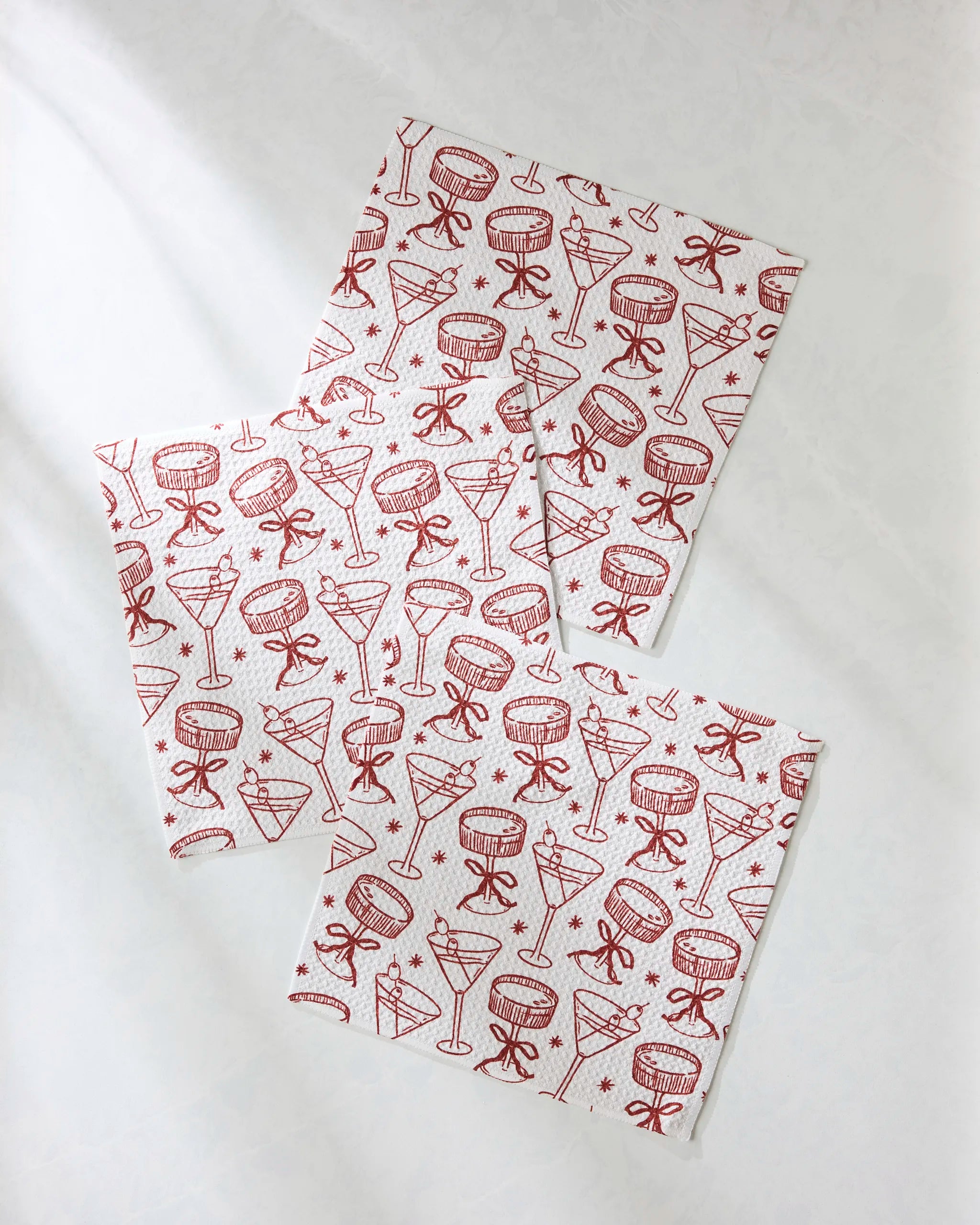 The Soiree set by Geometry features three white napkins made from recycled materials, adorned with red line drawings of festive cocktail glasses and bows—perfect as unique dishcloths or a charming decor accent for any gathering.