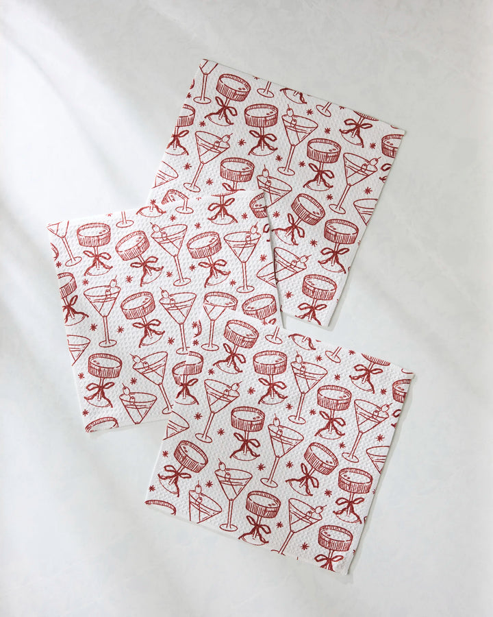 The Soiree set by Geometry features three white napkins made from recycled materials, adorned with red line drawings of festive cocktail glasses and bows—perfect as unique dishcloths or a charming decor accent for any gathering.