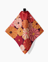 The Spicy Pumpkins napkin by Rebecca Bobko is a square fabric with a loop, showcasing vibrant stylized flowers in orange, red, pink, and yellow on a dark background—ideal for pairing with your favorite dishcloth set.