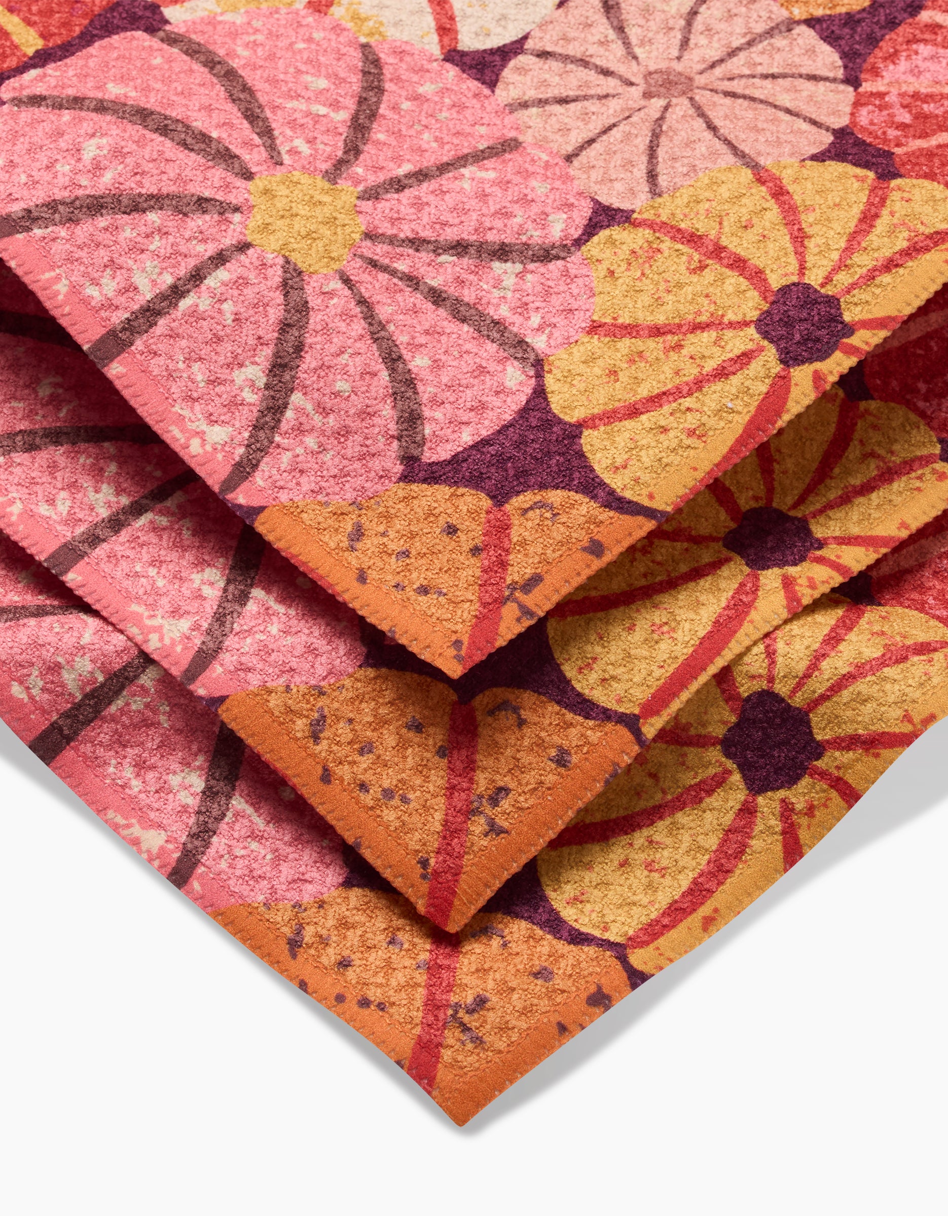 A close-up of the folded Spicy Pumpkins rug by Rebecca Bobko displays large, stylized flowers in pink, orange, yellow, and purple hues with textured details—evoking vibrant recycled dishcloth designs.