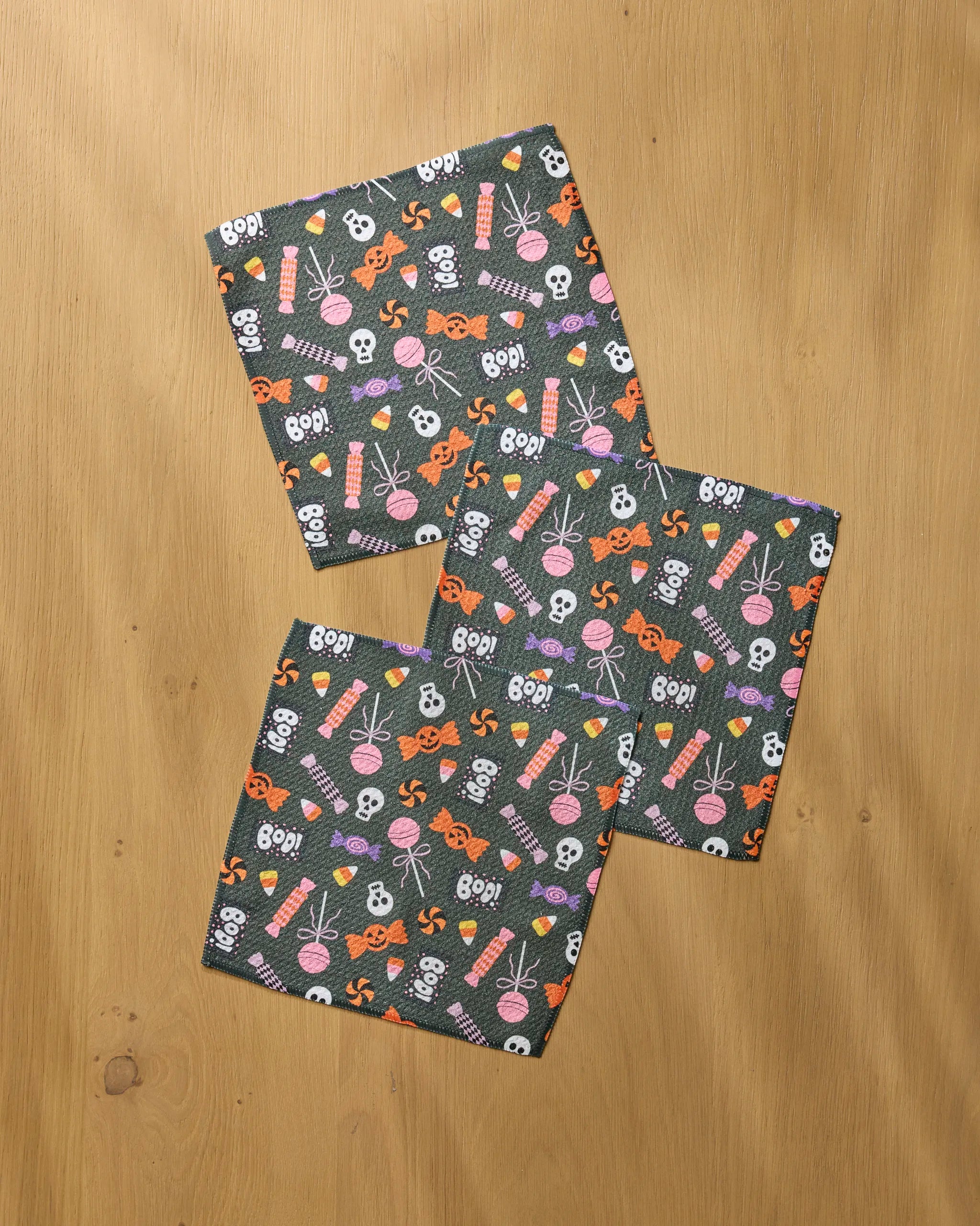 The Geometry Spooky Sweets Black set includes three Halloween-themed recycled dishcloths with skulls, spider webs, candy, pumpkins, and "BOO!" in orange, white, and pink, artfully arranged on a light wooden surface.