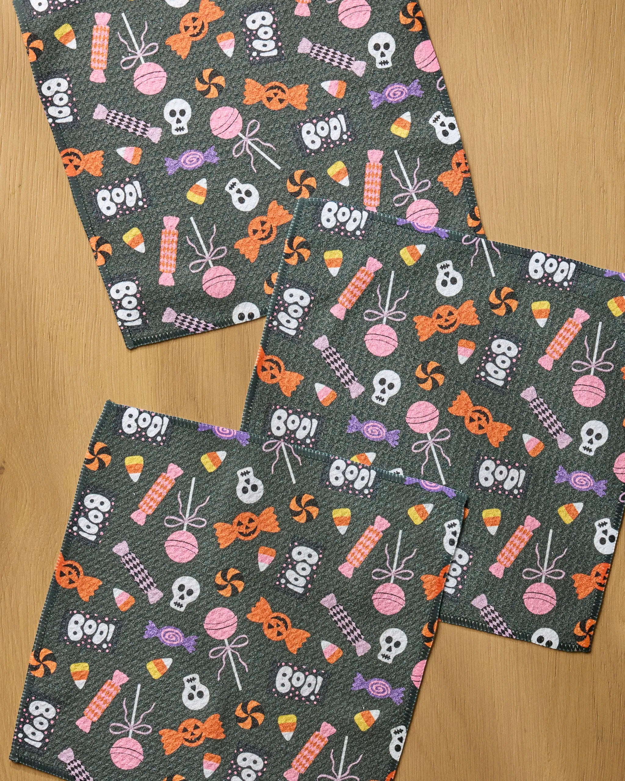 Three absorbent dark gray napkins from Geometry's Spooky Sweets Black set, featuring Halloween skulls, bones, spiders, candies, and "BOO!", add festive fun to your kitchen décor.