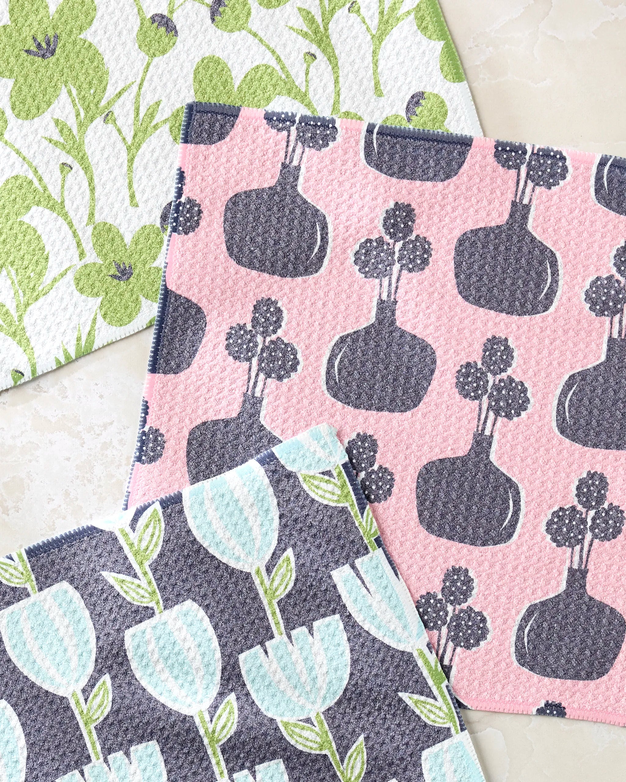 Lizzie Clark’s Spring Bouquet set features three patterned dishcloths: one with green flowers, one with blue tulips and green stems, and one with gray vases and round flowers on a pink background. All are made from recycled materials.