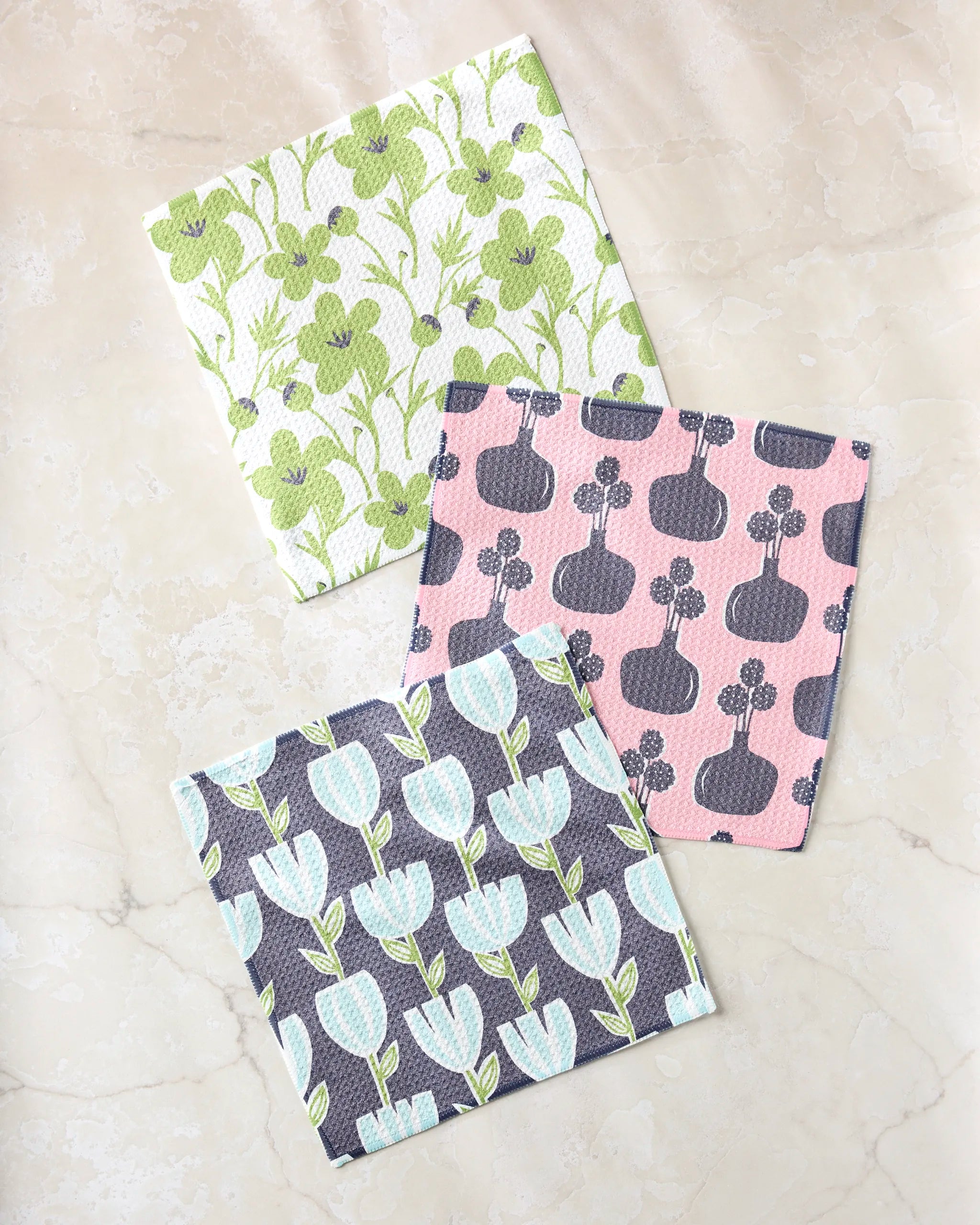 Three Lizzie Clark Spring Bouquet cloths made from recycled materials are displayed on light marble: one with green florals, one with pink and gray pumpkins, and a third featuring blue and white tulips on gray.
