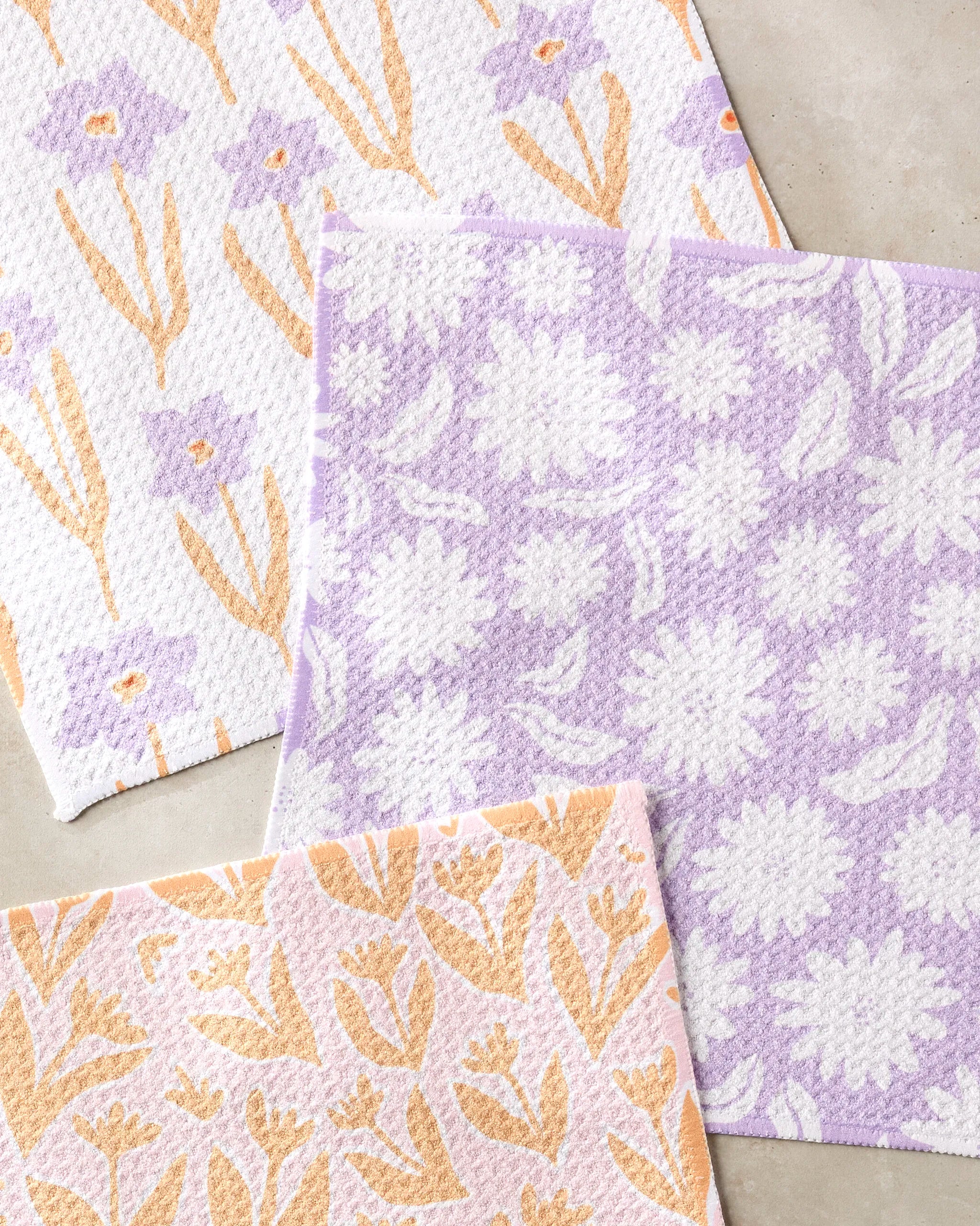 Three overlapping "Spring Flowers" dishcloths by Kayla Ann are shown on a light surface, each super-absorbent white cloth featuring pastel purple and yellow botanical patterns.