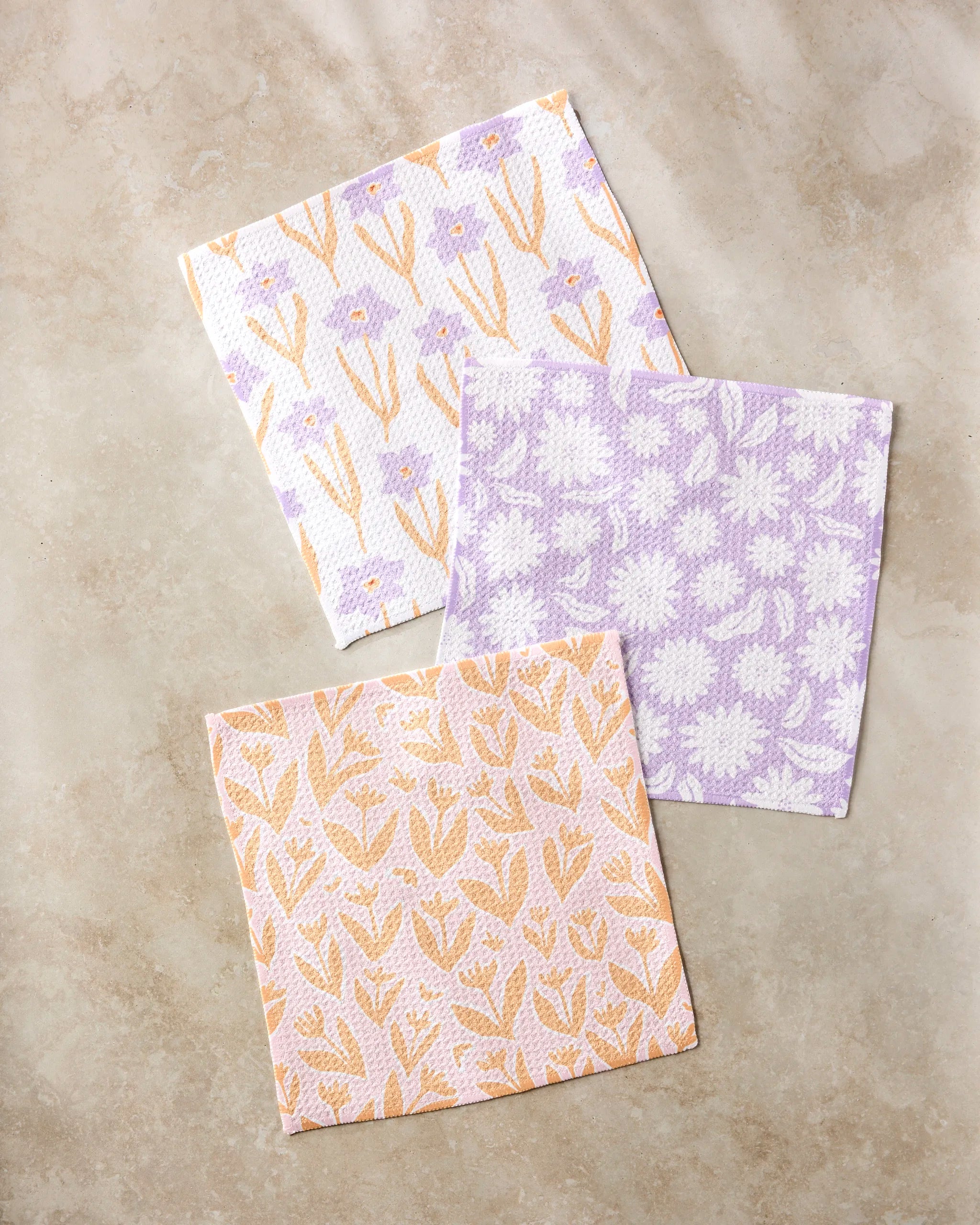 The Kayla Ann Spring Flowers set features three square, super-absorbent dishcloths in lavender, peach, and white, each with a unique floral pattern, beautifully displayed on a beige marble surface.