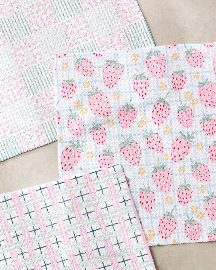 The Spring Picnic Day dishcloth set by Hufton Studio includes three super-absorbent cloths: pink strawberries with yellow flowers, a pink and green plaid, and pink stripes with black X patterns—all made from recycled materials.