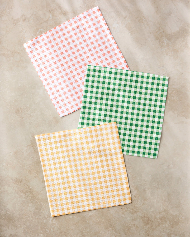 Three Spring Plaid napkins by Danelys Sidron, made from recycled materials, are displayed on a light beige surface. The set includes pink and white, green and white, and yellow and white gingham checkered patterns.