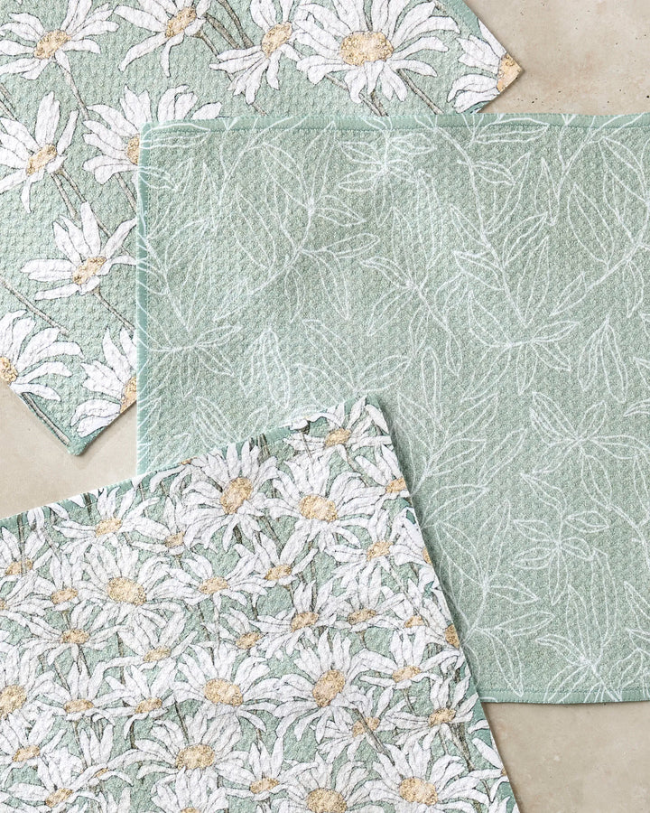 Three textured green cloths from the Aubrey Fairchild "Springtime" recycled kitchen towel set are arranged on a light beige surface—two with daisies and one with white leaves.