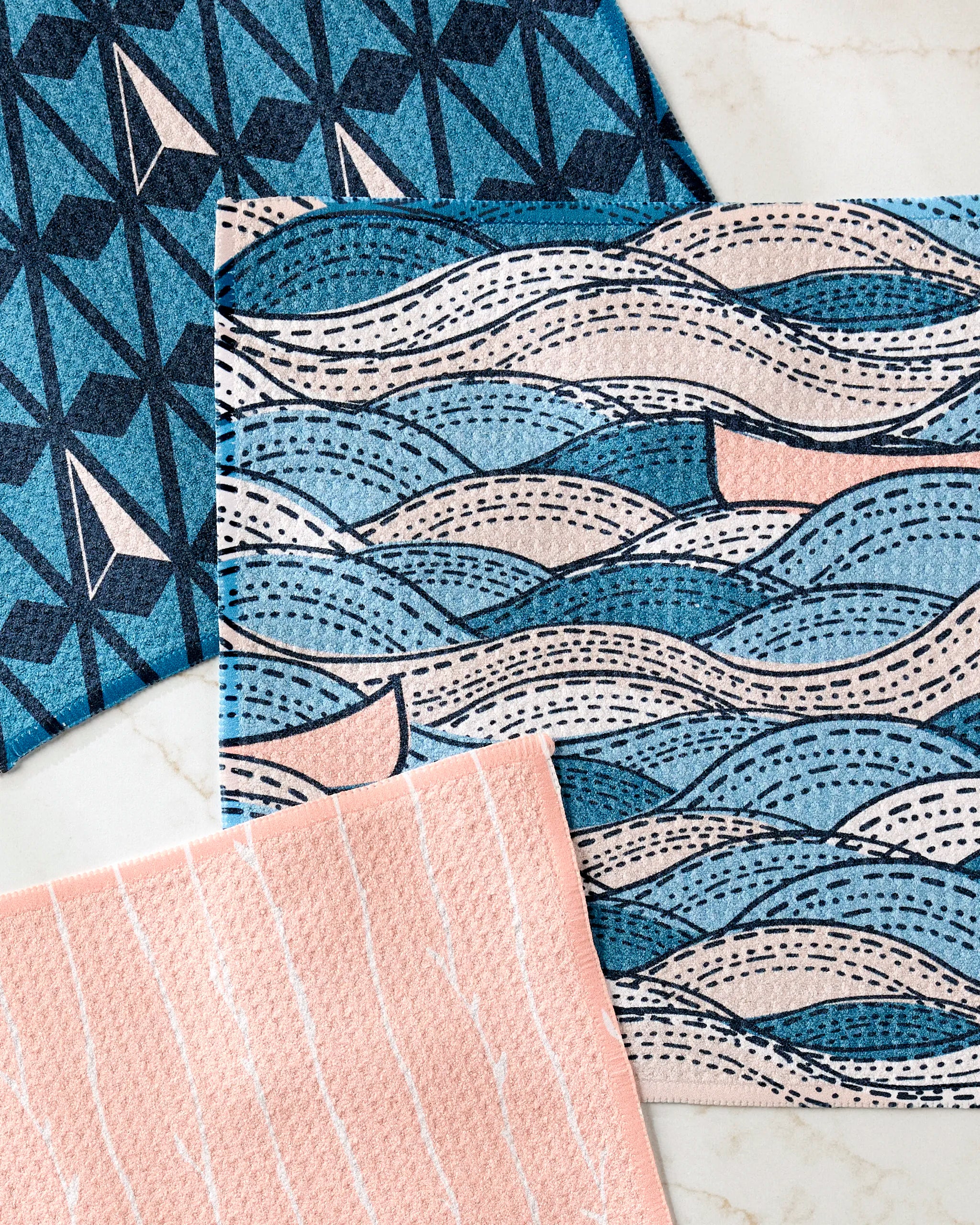 Three rectangular Twig + Spark Squall super-absorbent dishtowels, featuring blue and pink geometric and wavy patterns, are laid out slightly overlapping on a light-colored marble surface.